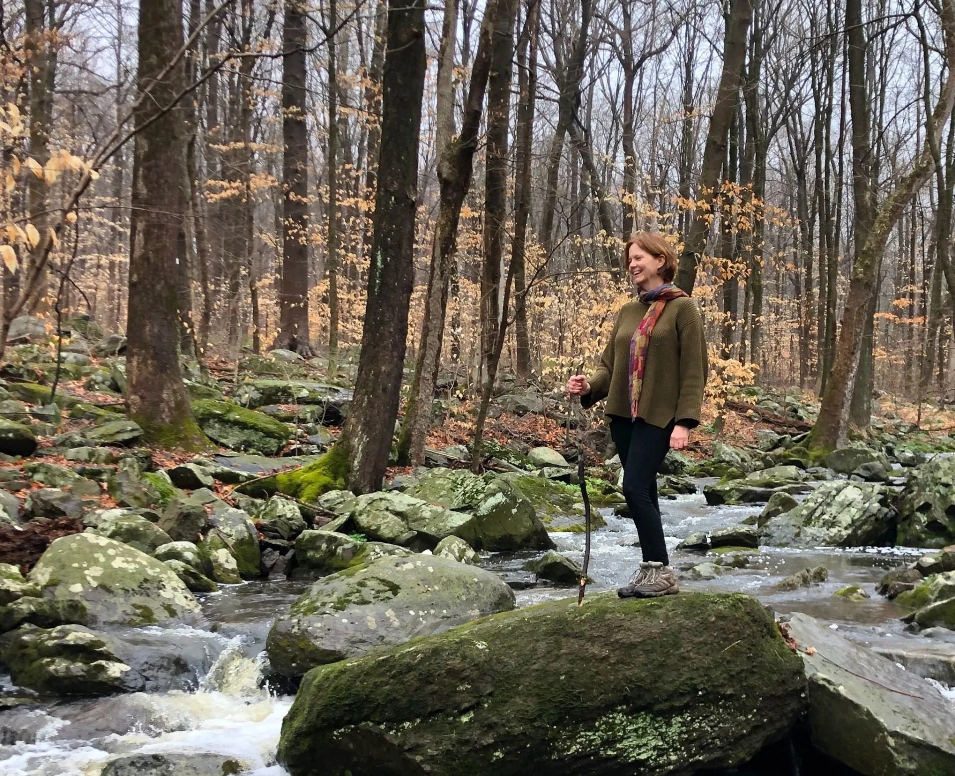 A woman standing on a large mossy rock in a shallow stream in a forest during autumn. She is smiling, holding a walking stick, and wearing hiking shoes, a green sweater, and a scarf. The trees in the background have mostly bare branches and some brown leaves.