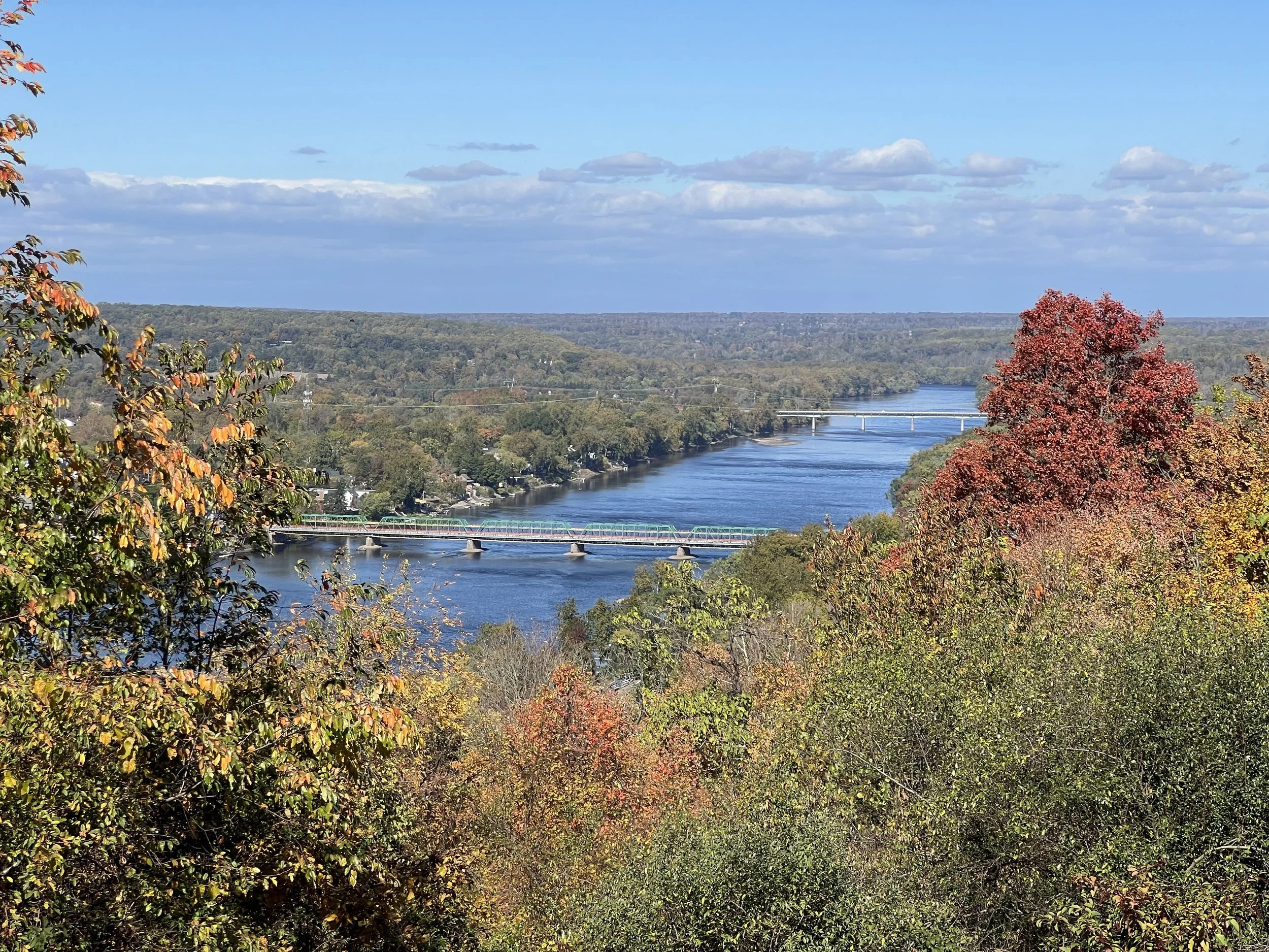 Scenic view of a river flowing through a hilly landscape with colorful trees in fall foliage, bridges crossing the river, and a distant horizon with blue sky and clouds.