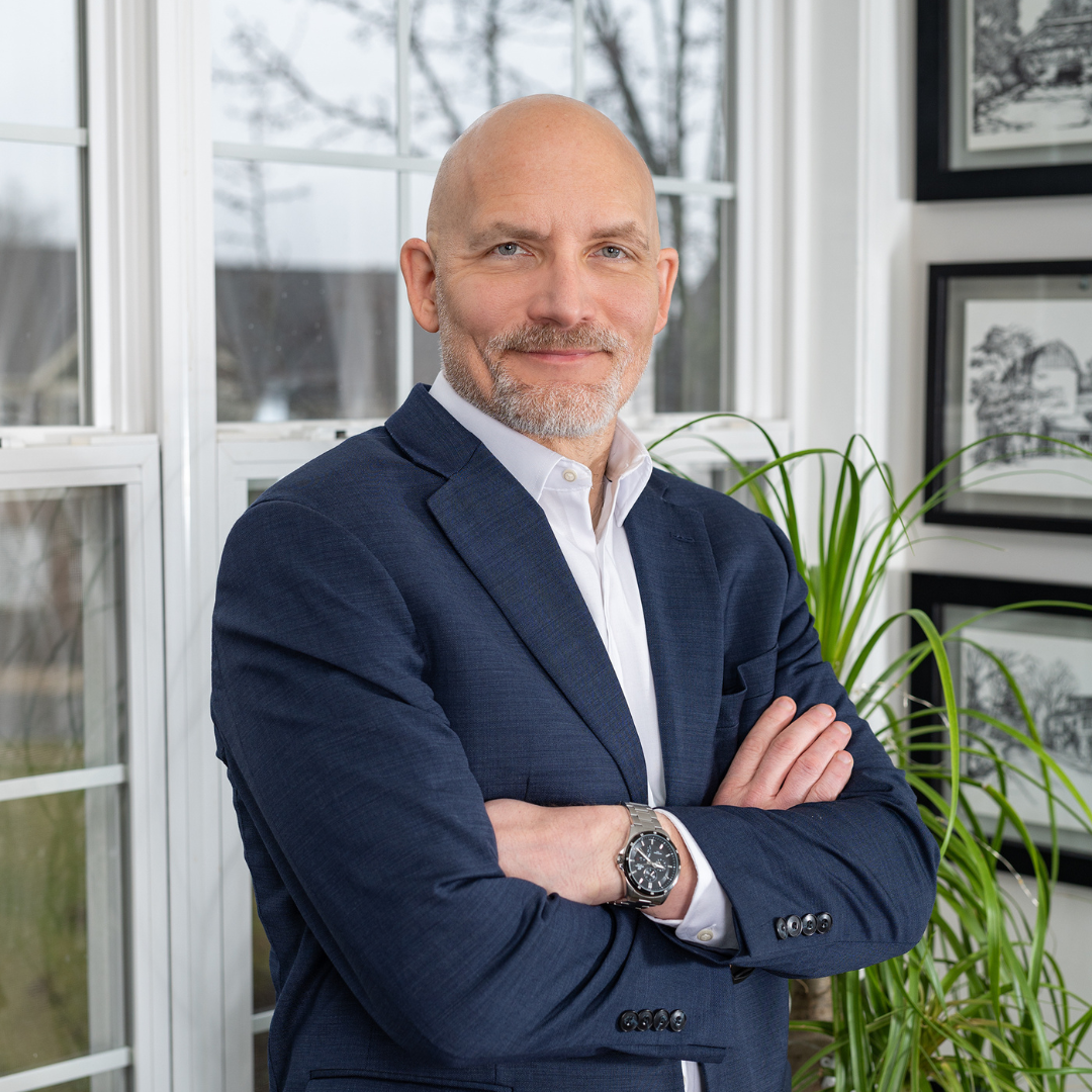 A middle-aged man with a bald head and a beard, wearing a navy blue suit and white shirt, stands with arms crossed in a bright room with large windows and framed black and white pictures on the walls.