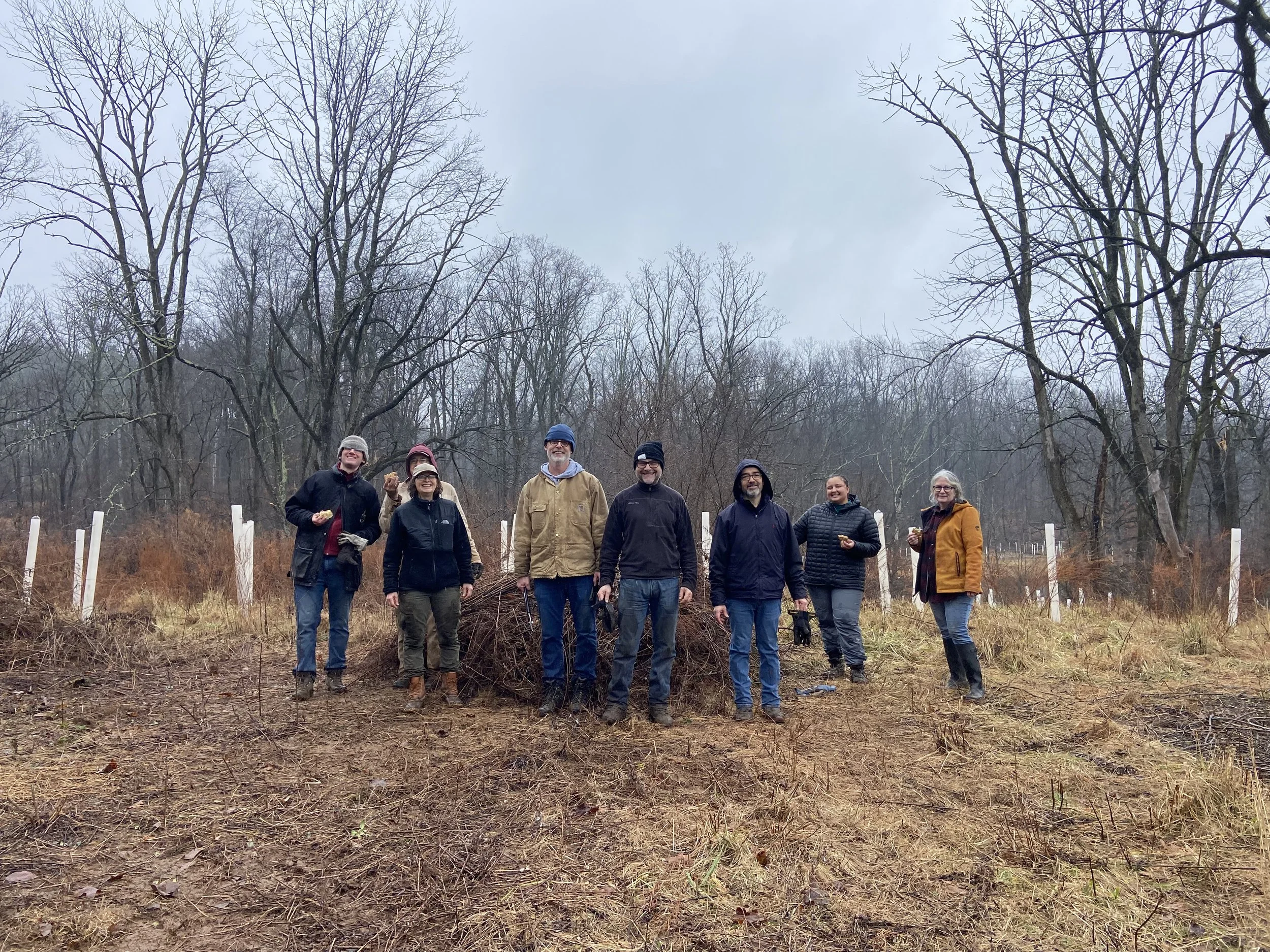 Group of seven people standing outdoors in a wooded area during a cloudy day, some holding small objects, with sapling trees planted behind them.