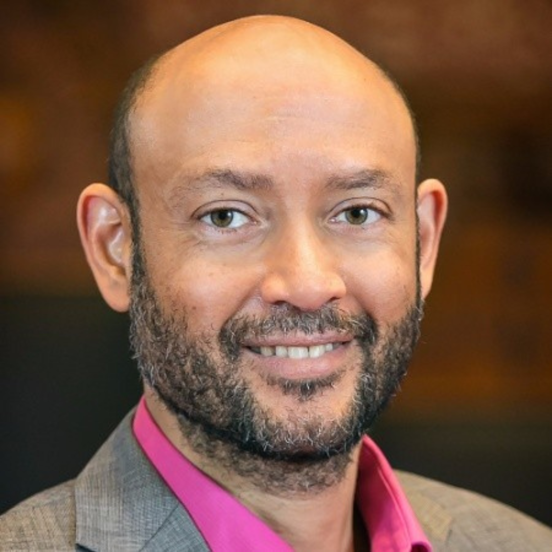 Close-up portrait of a smiling man with a beard, light skin, and wearing a pink shirt and a grey blazer.