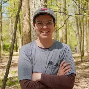 A smiling young person wearing glasses and a baseball cap, standing with arms crossed in a wooded area during the daytime.