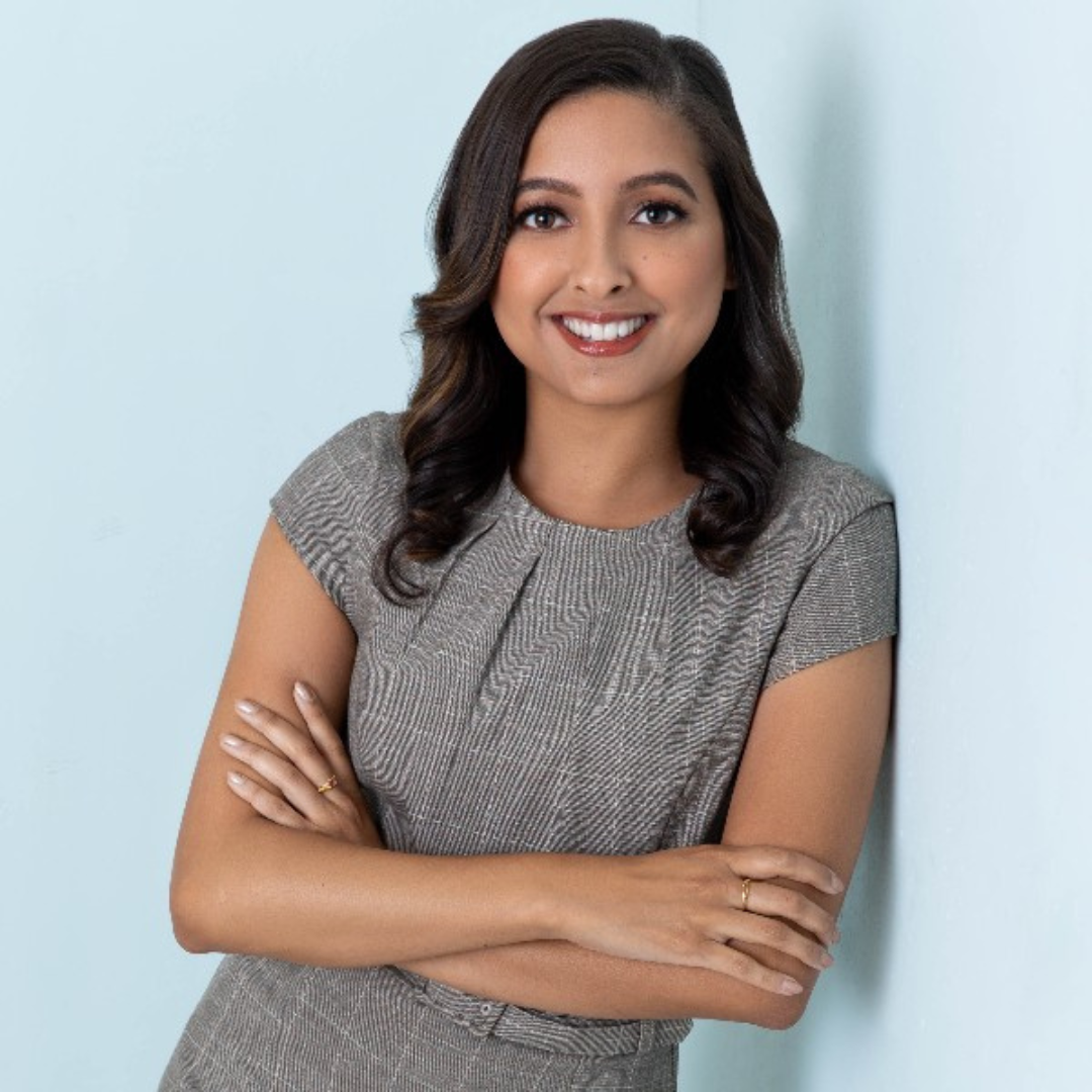 A woman with dark brown hair, wearing a gray short-sleeved top, smiling, standing against a light blue wall.