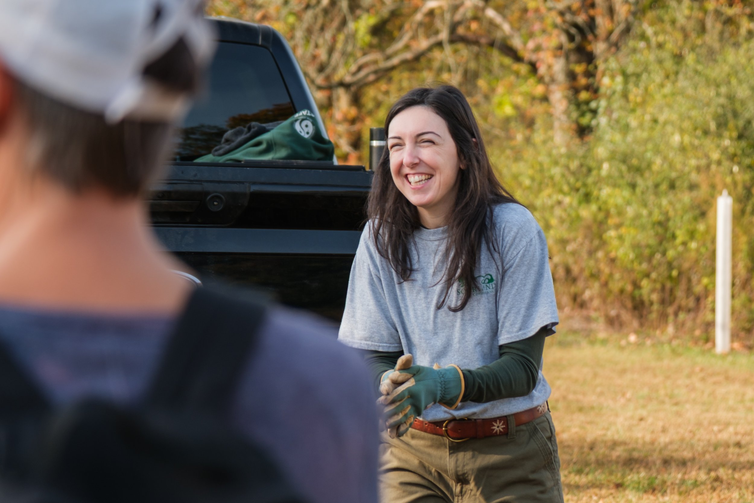 A woman with long dark hair smiling outdoors in front of a black pickup truck, wearing a gray t-shirt, green long sleeve shirt, and gloves, with autumn trees in the background.