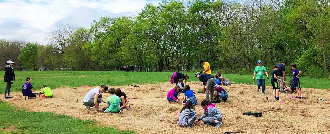 Children and adults planting trees in a field during daytime under cloudy sky, with green trees in the background.