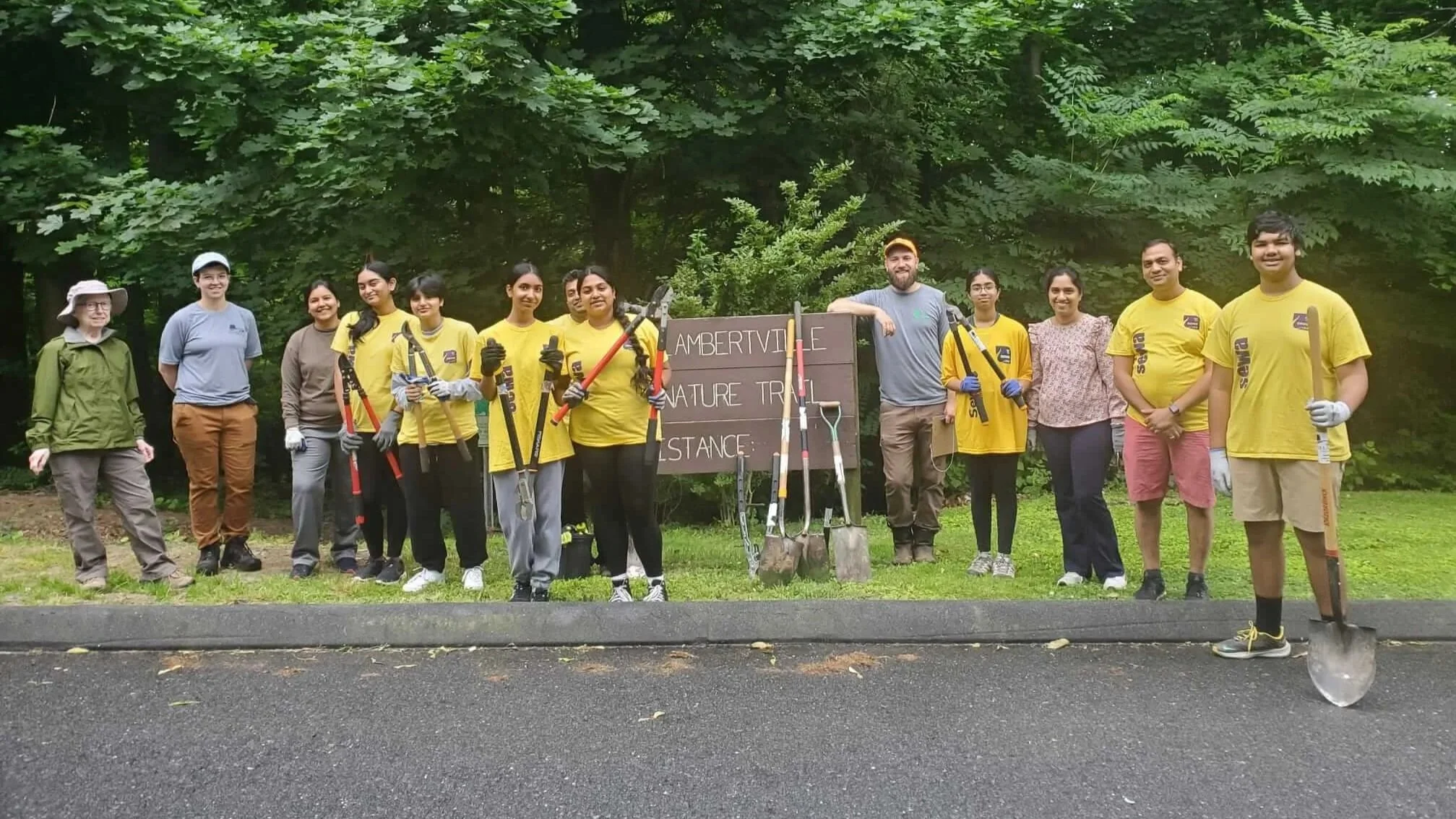 Group of volunteers at Ambertville Nature Trail with tools like shovels, rakes, and pruning shears, standing outdoors in front of a sign and trees, wearing gloves and casual clothing.