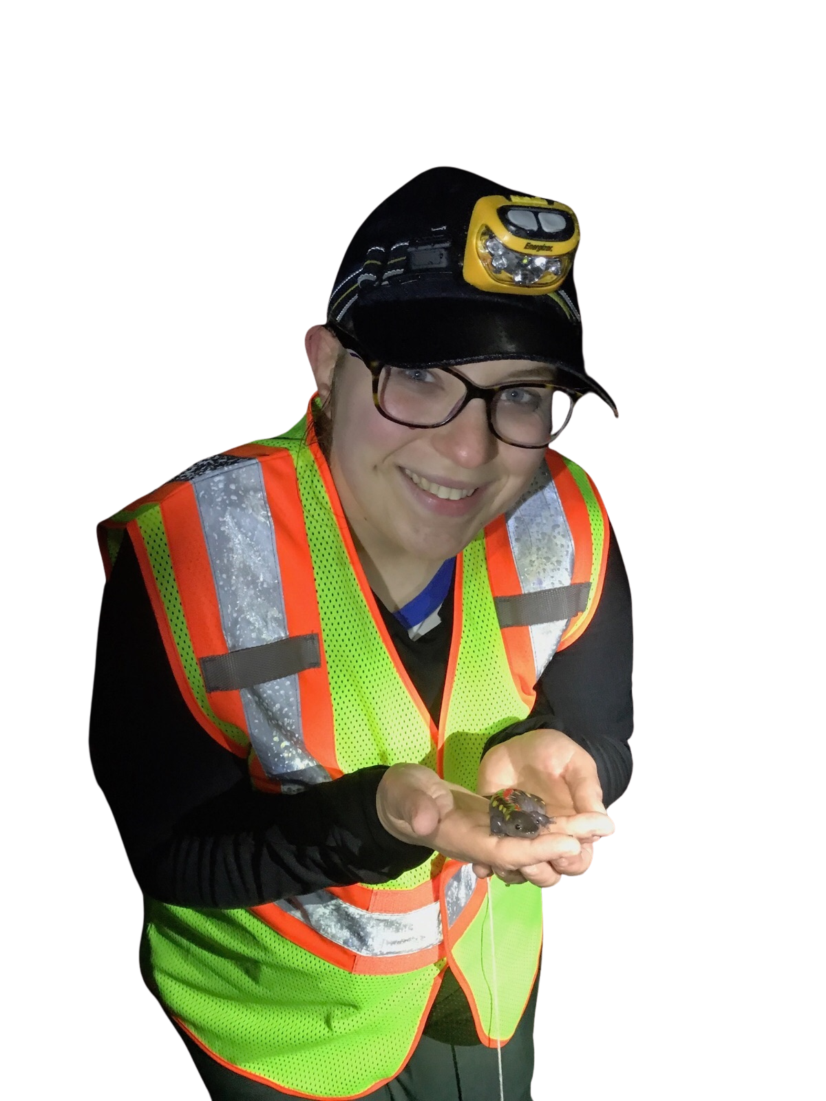 A smiling woman wearing glasses, a reflective safety vest, and a headlamp on her black cap, holding a small insect in her hands against a dark background.