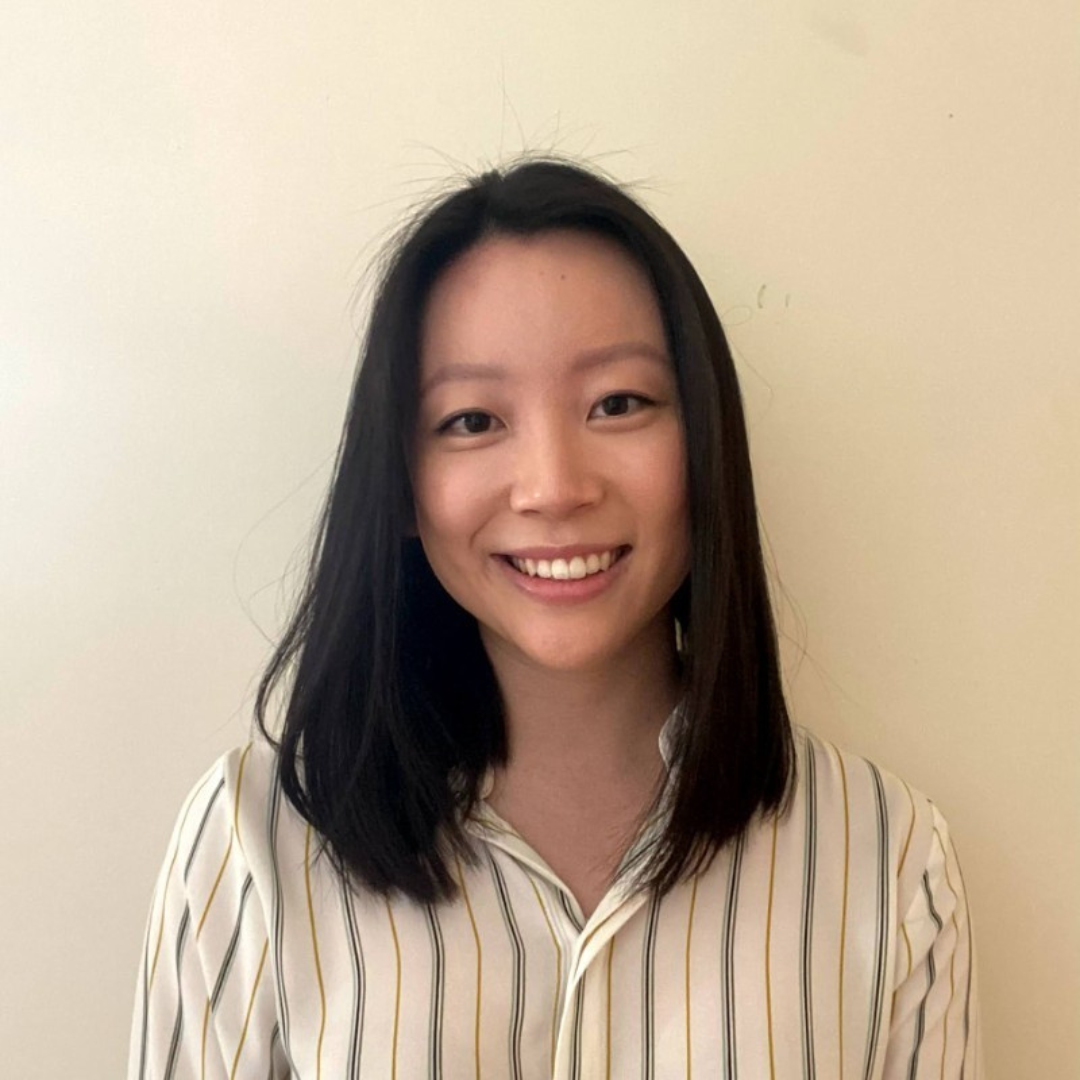 A young woman with shoulder-length black hair smiling at the camera, wearing a beige and white striped shirt, standing against a plain light-colored background.