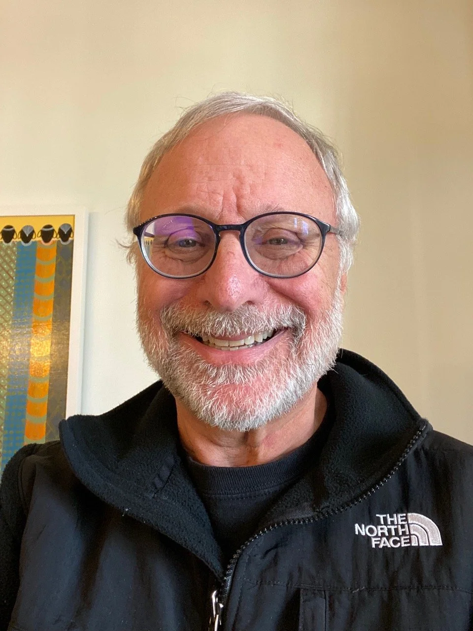 A smiling elderly man with glasses, a gray beard, and gray hair, wearing a black North Face jacket, indoors with a beige wall and a colorful abstract painting in the background.