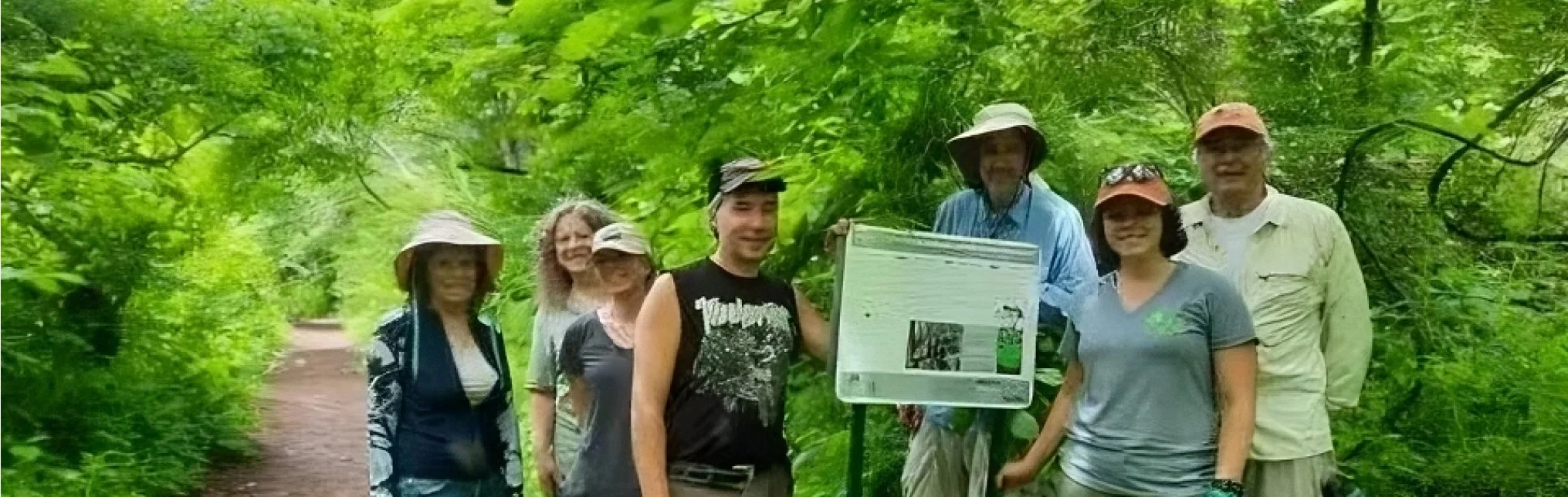 Group of seven people standing on a trail in a lush green forest, some wearing hats, near an informational sign.