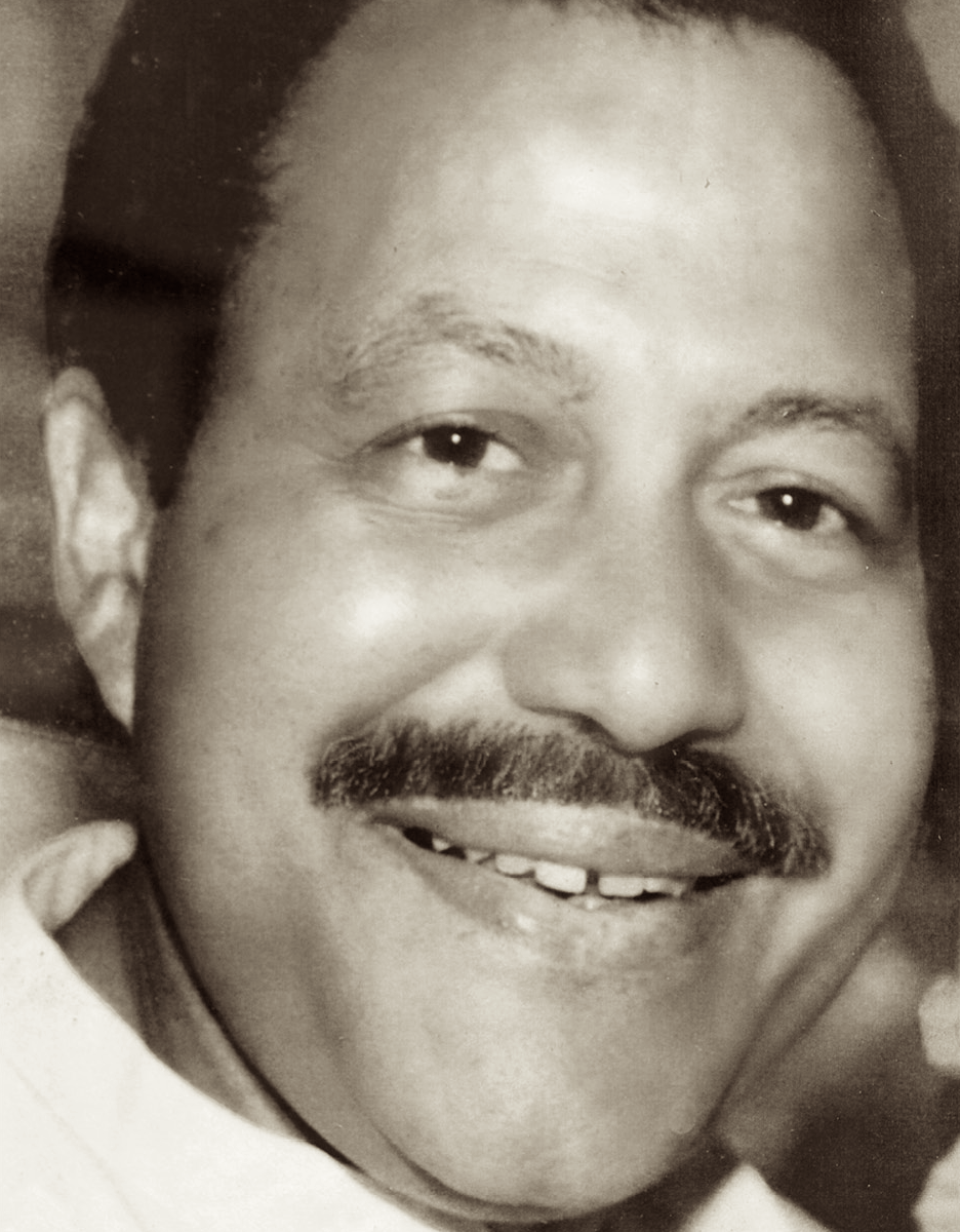 A black-and-white photo of a man with a mustache, smiling, with short hair, and missing a tooth.