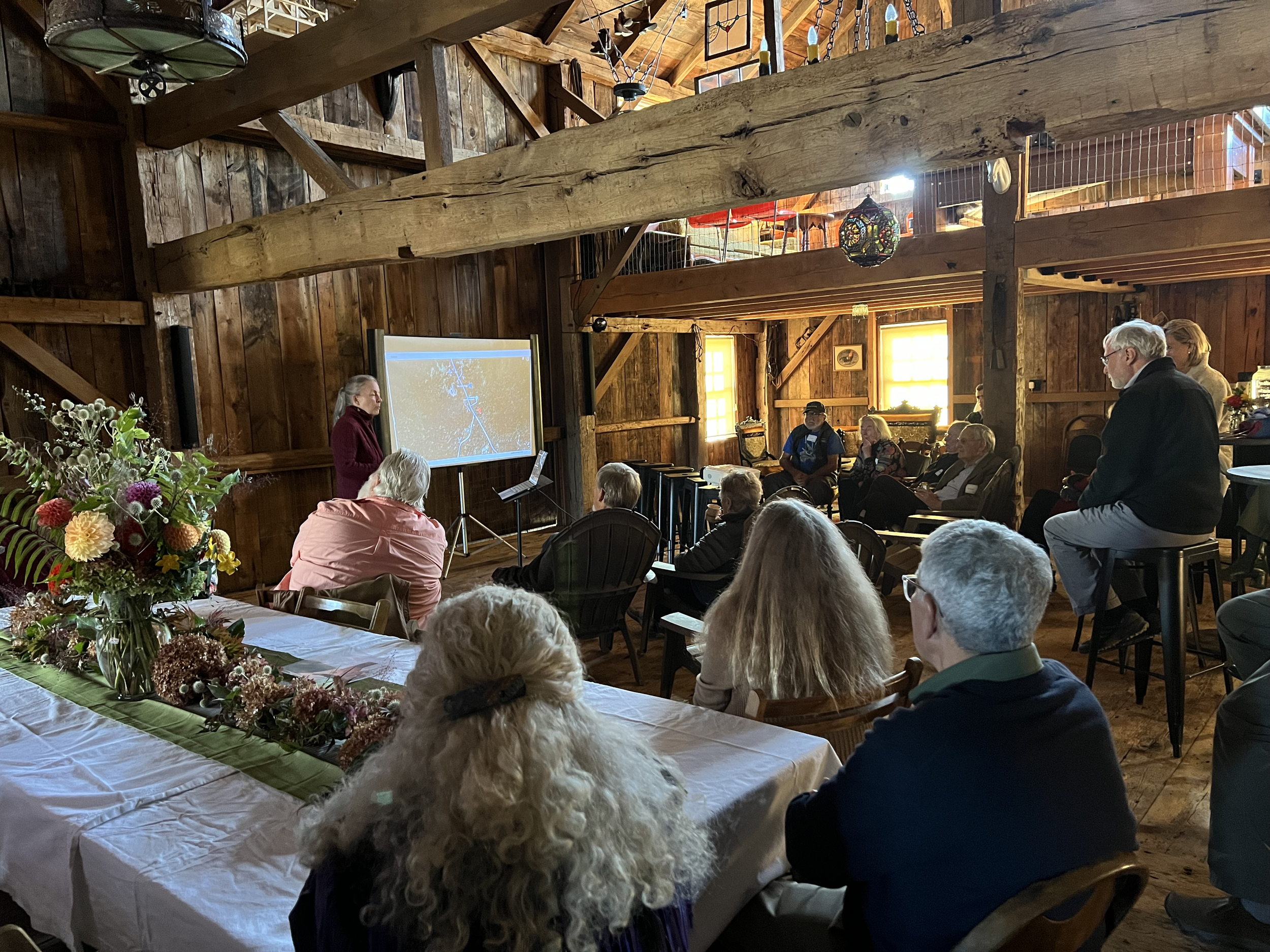 A group of people attending a presentation in a rustic wooden barn-like setting, with a woman speaking at a screen and a table with a floral centerpiece in the foreground.