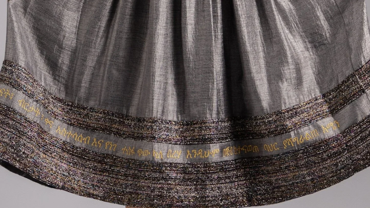 Close-up of Amharic text embroidery along the hem of a grey pleated midi skirt.