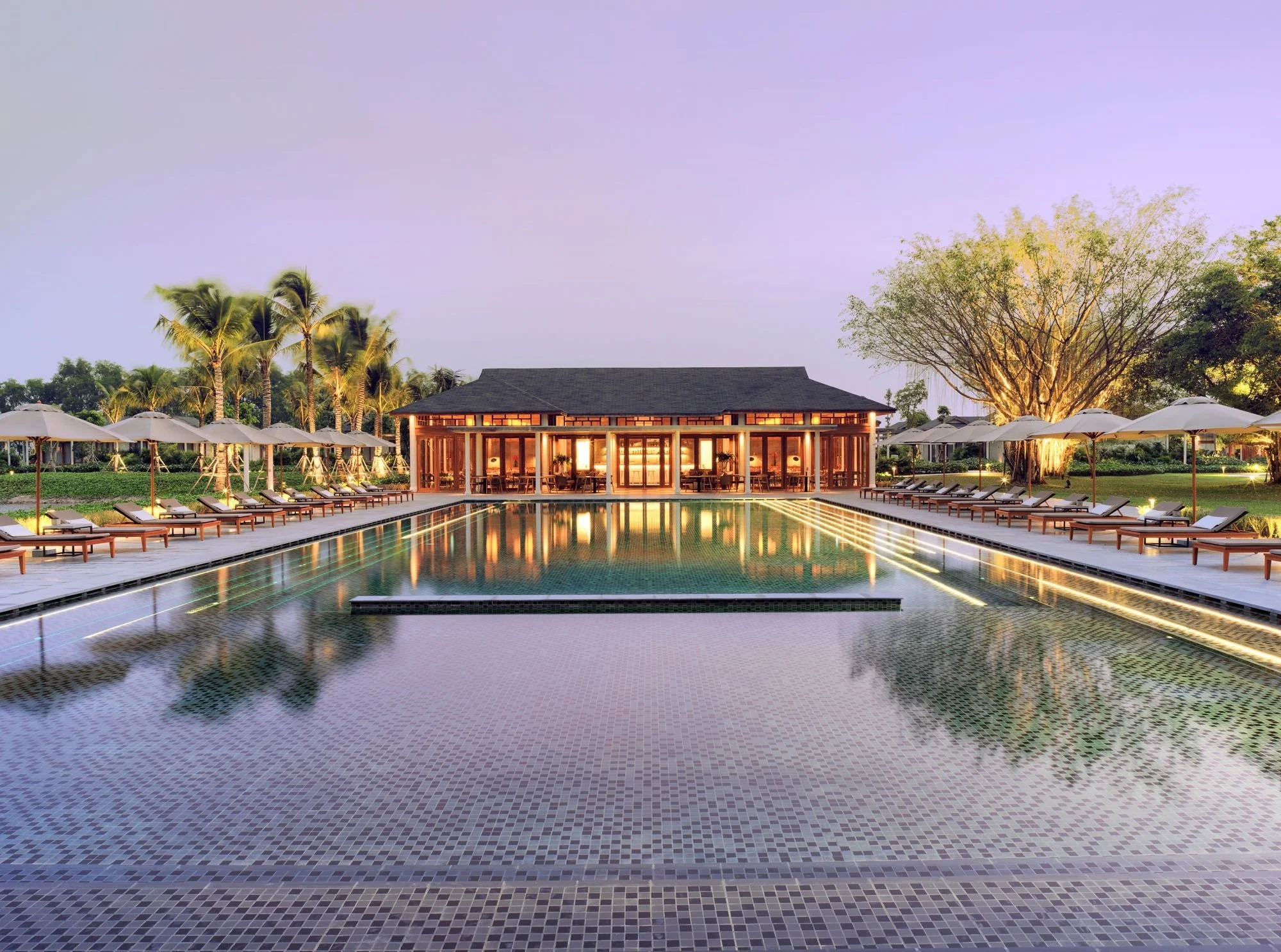 Luxury resort pool in Can Tho, Vietnam, with sun loungers, umbrellas, tropical landscaping, and a softly lit pavilion at dusk.