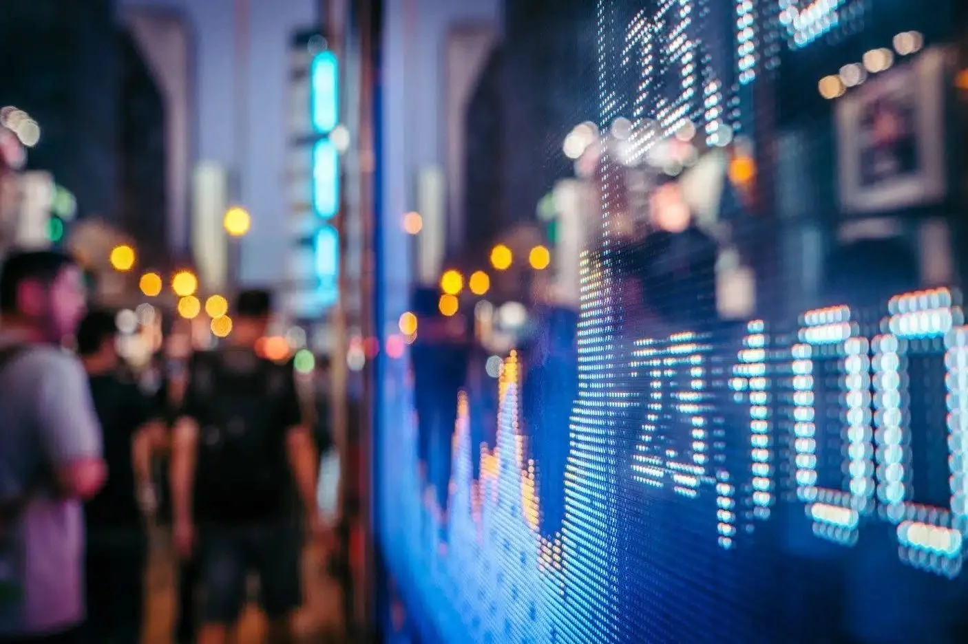 A digital stock ticker display glowing with data visuals, reflected in a busy city street filled with blurred silhouettes.