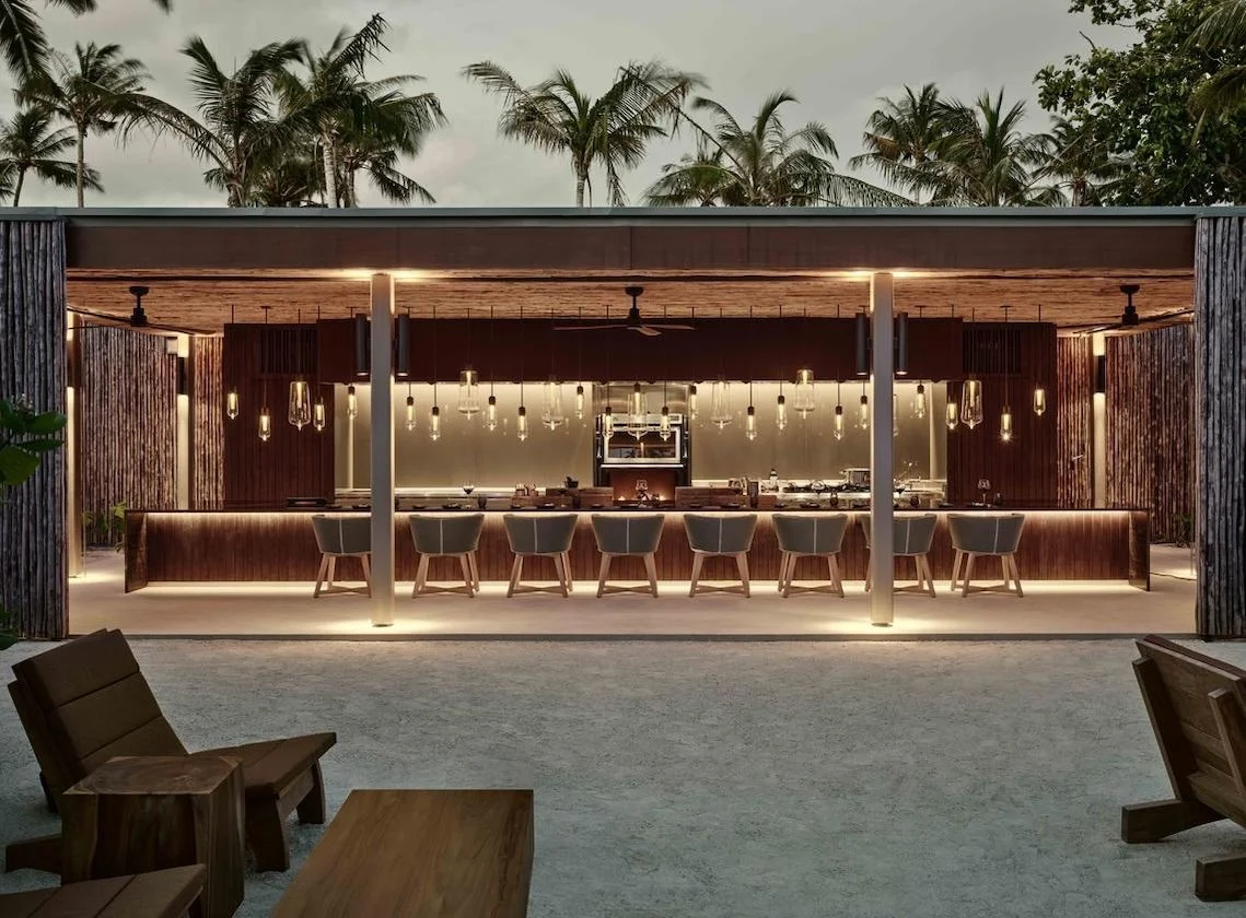 Open-air beachfront bar glowing at dusk, framed by palms and minimalist wooden design.