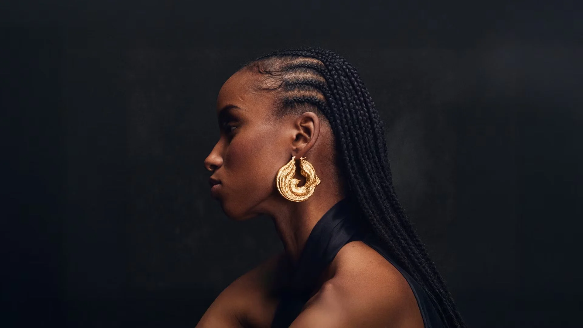 Side profile of a model with braided hair wearing bold sculptural gold earrings, against a dark backdrop, showcasing luxury accessory styling.