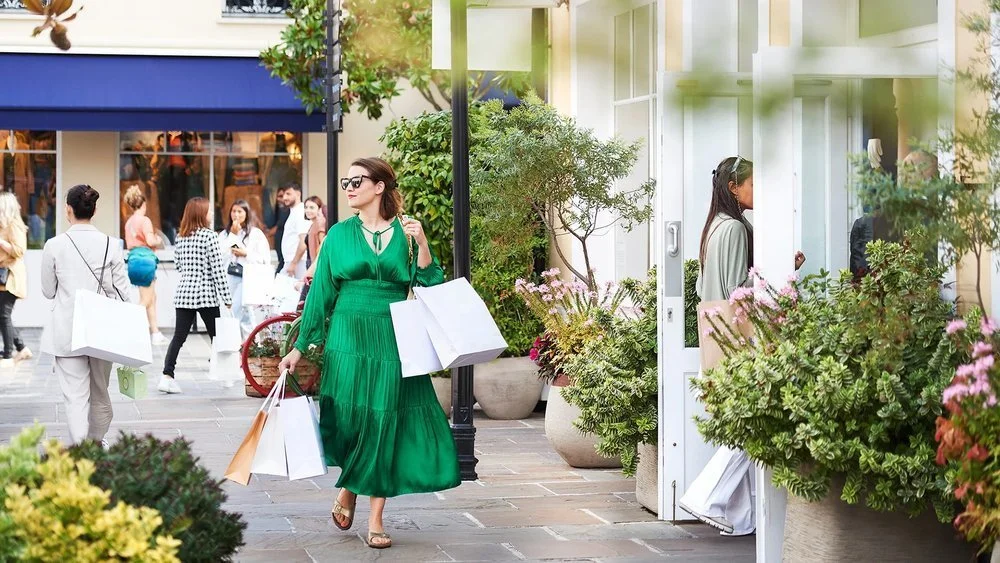 A woman in a green dress walks with shopping bags at a La Valle Village in Paris, showcasing high-end retail lifestyle.
