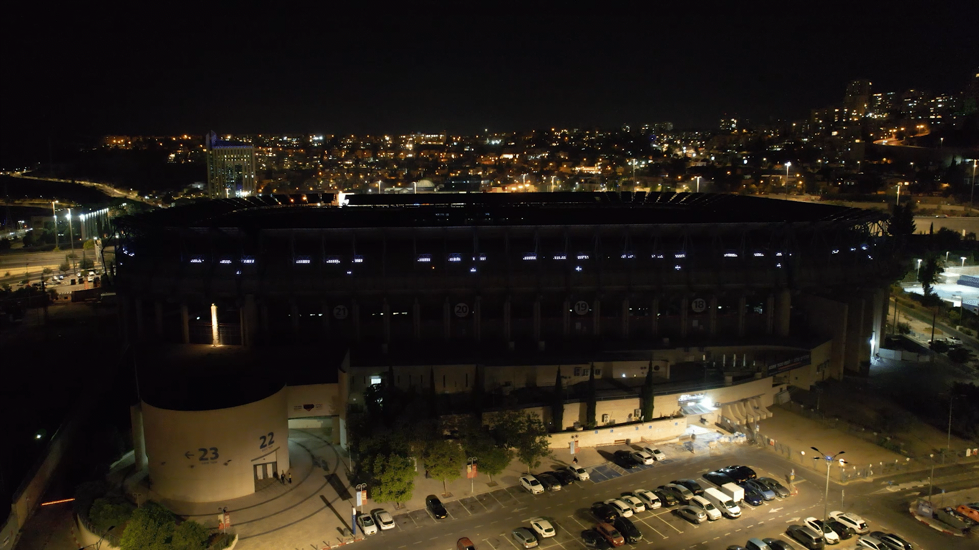 Nighttime aerial view of a large illuminated stadium with a city skyline in the background, parking area with cars in the foreground.