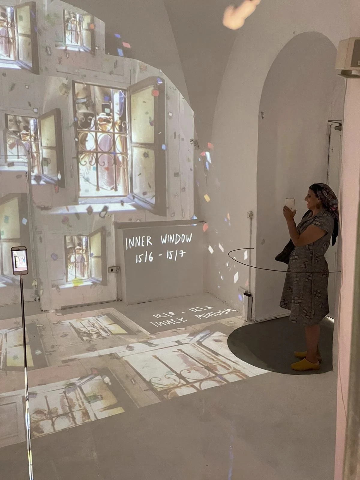 A woman standing on a black circular mat indoors, taking a photo with her phone. The floor and wall are covered with a video projection of windows, including an open window with a view outside. The projection shows various window views with iron bars