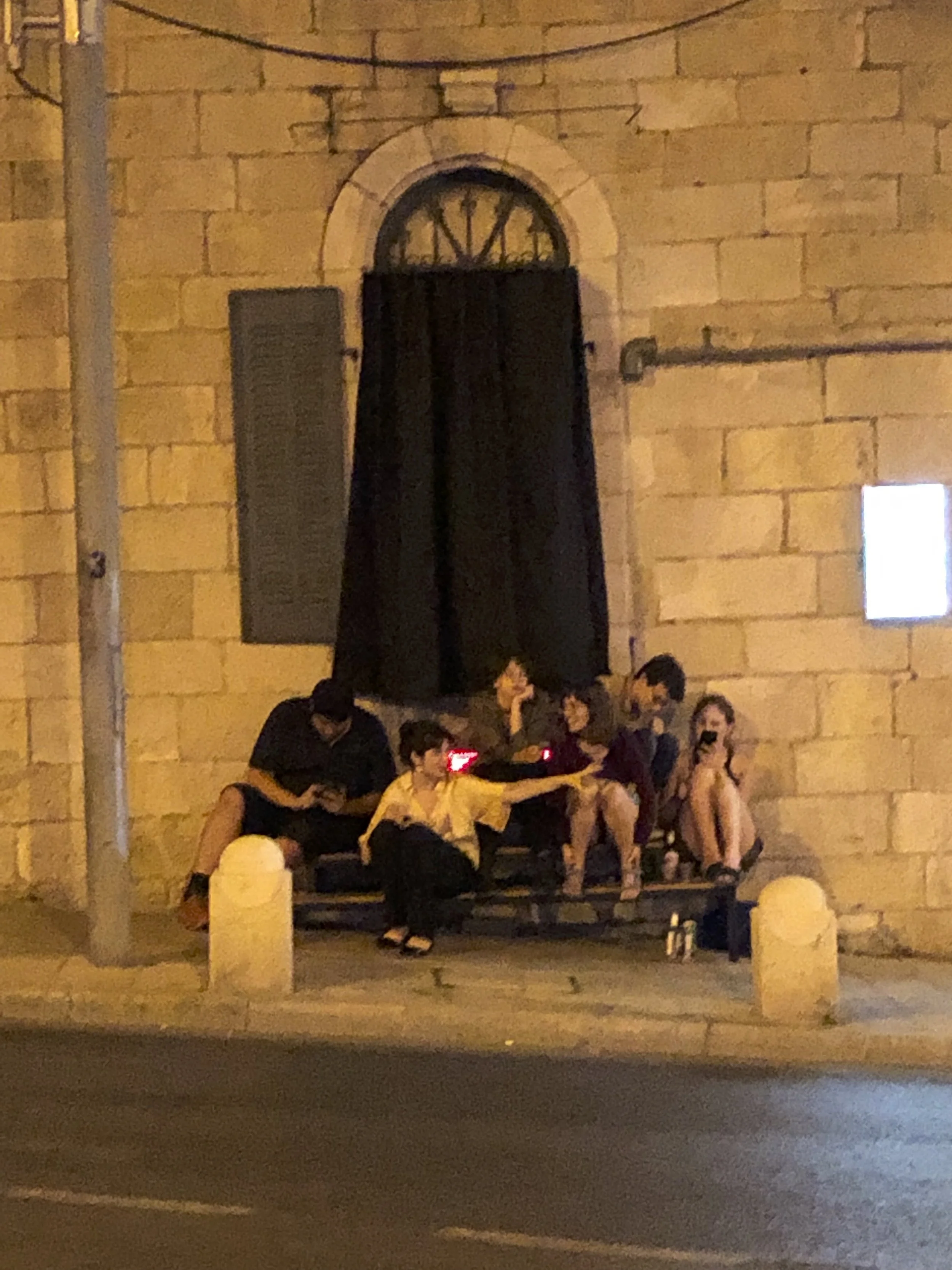 Group of five people sitting and standing on a bench outside a stone building at night. One person is using a smartphone, and others are interacting, with one holding an object. The scene is illuminated by streetlights.