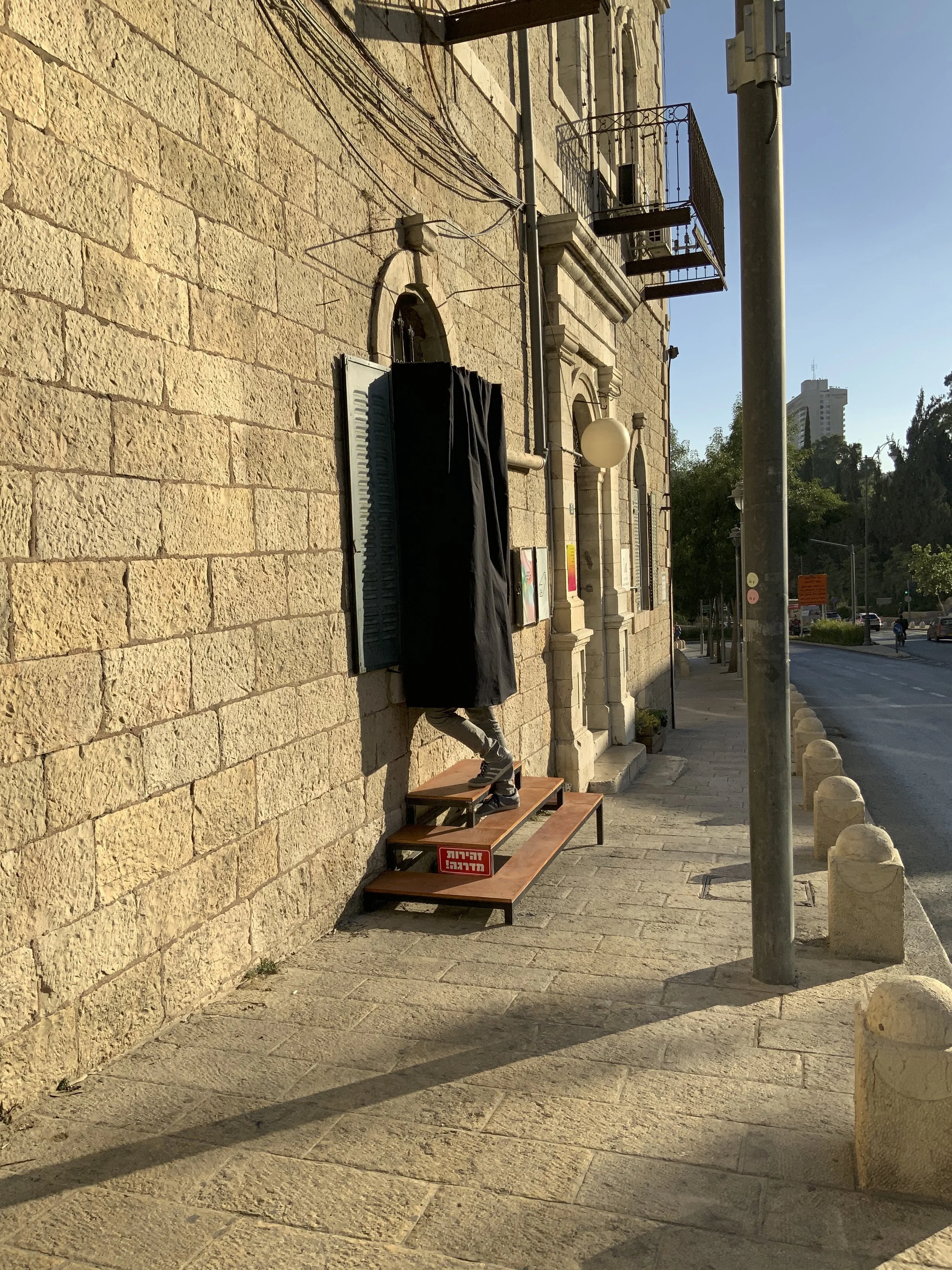 A person walking up a wooden bench outside a stone building, with part of their legs and black clothing visible, as they step onto the bench in front of a shuttered window.