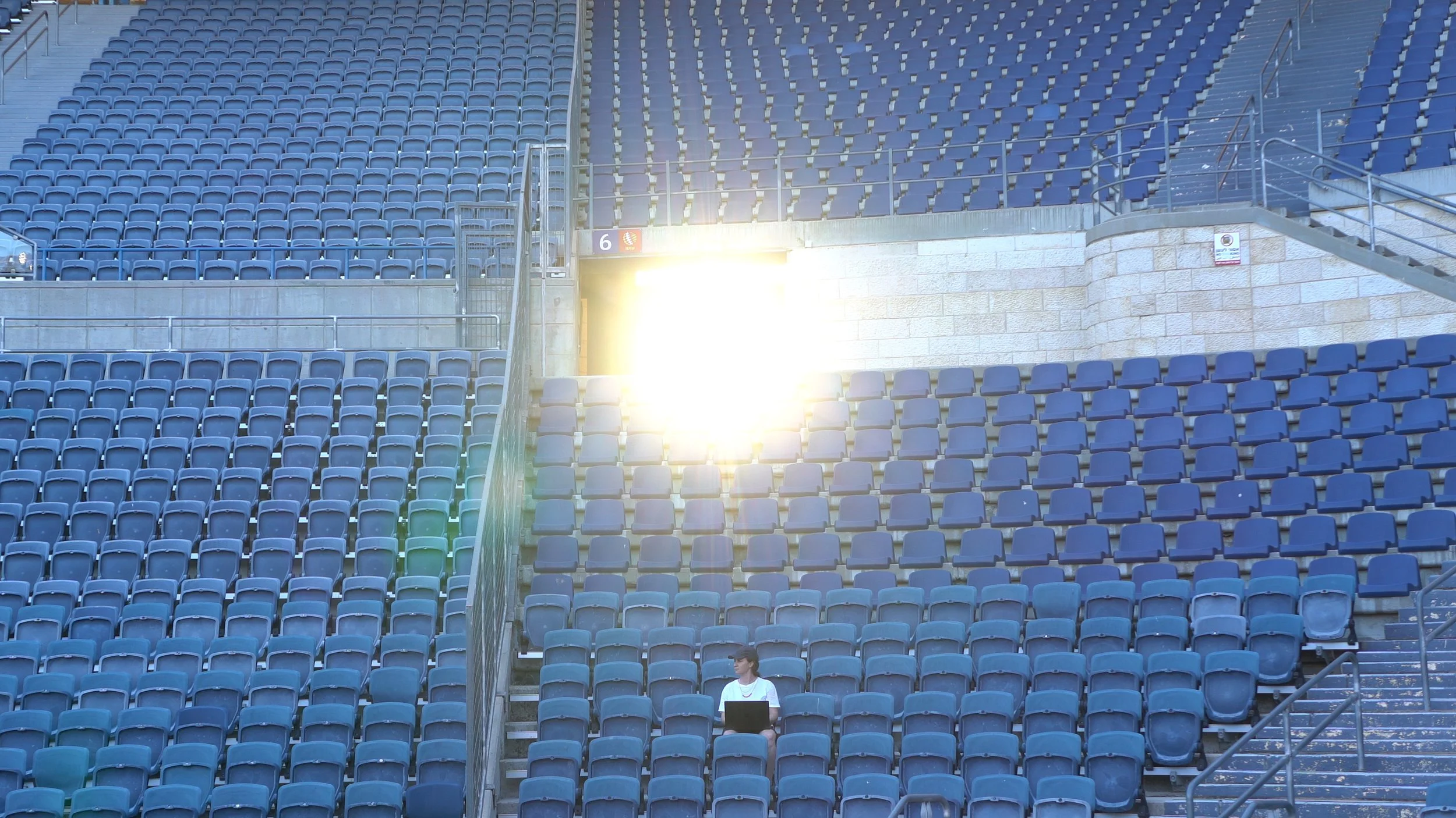 A young person sitting alone in an empty stadium with many blue seats, using a laptop on their lap, near a bright light shining on the seats.