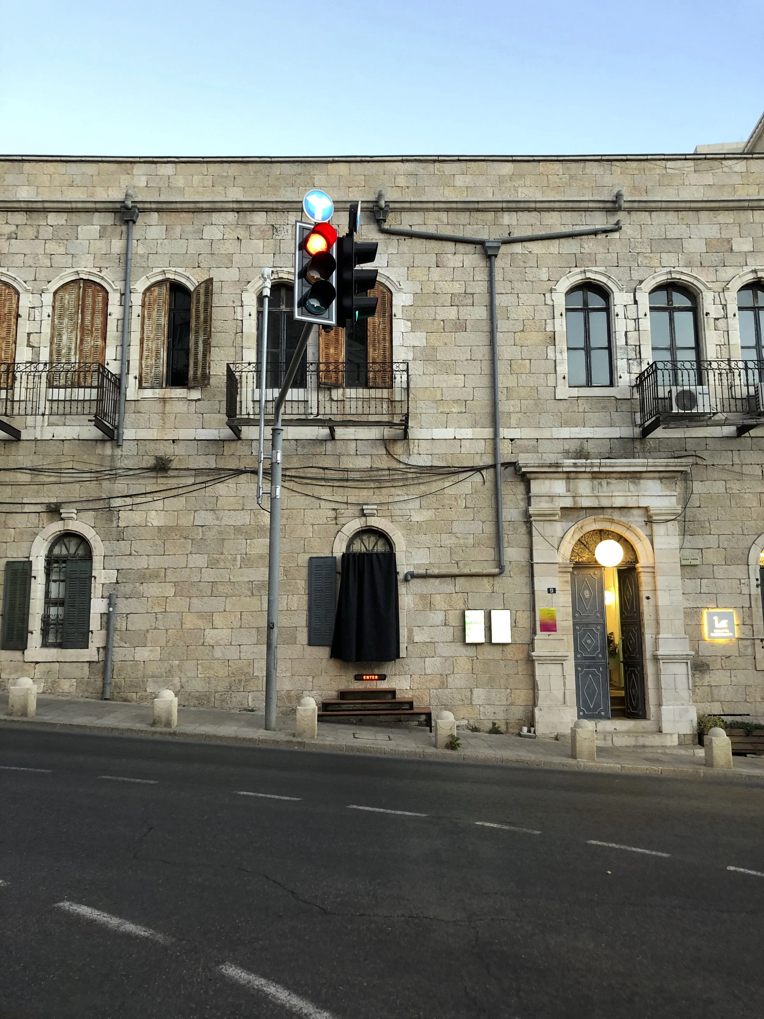 A street scene with a traffic light showing red, a stone building with arched windows, closed shutters, small balconies, a door with a lit round lamp above, and some signage. Urban setting.
