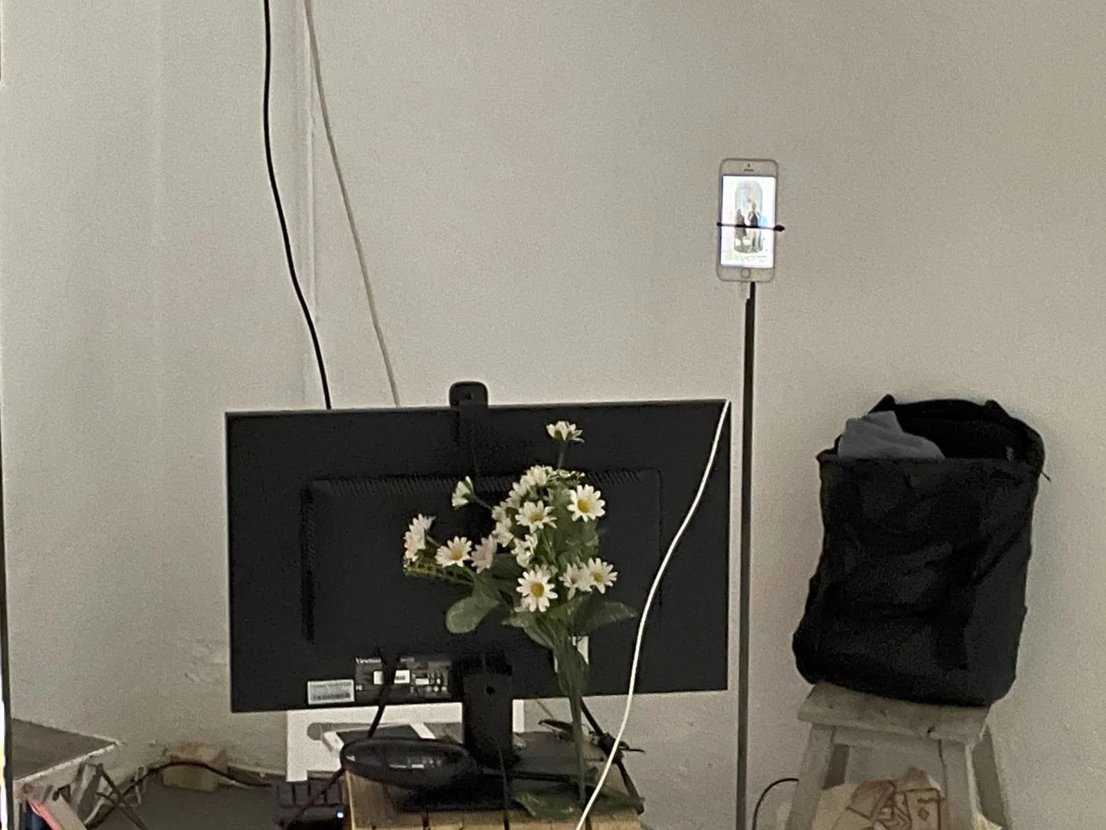 A computer monitor on a desk with a flower arrangement in front, a phone on a stand in the background capturing a picture, and a black bag on a stool beside a plain wall.