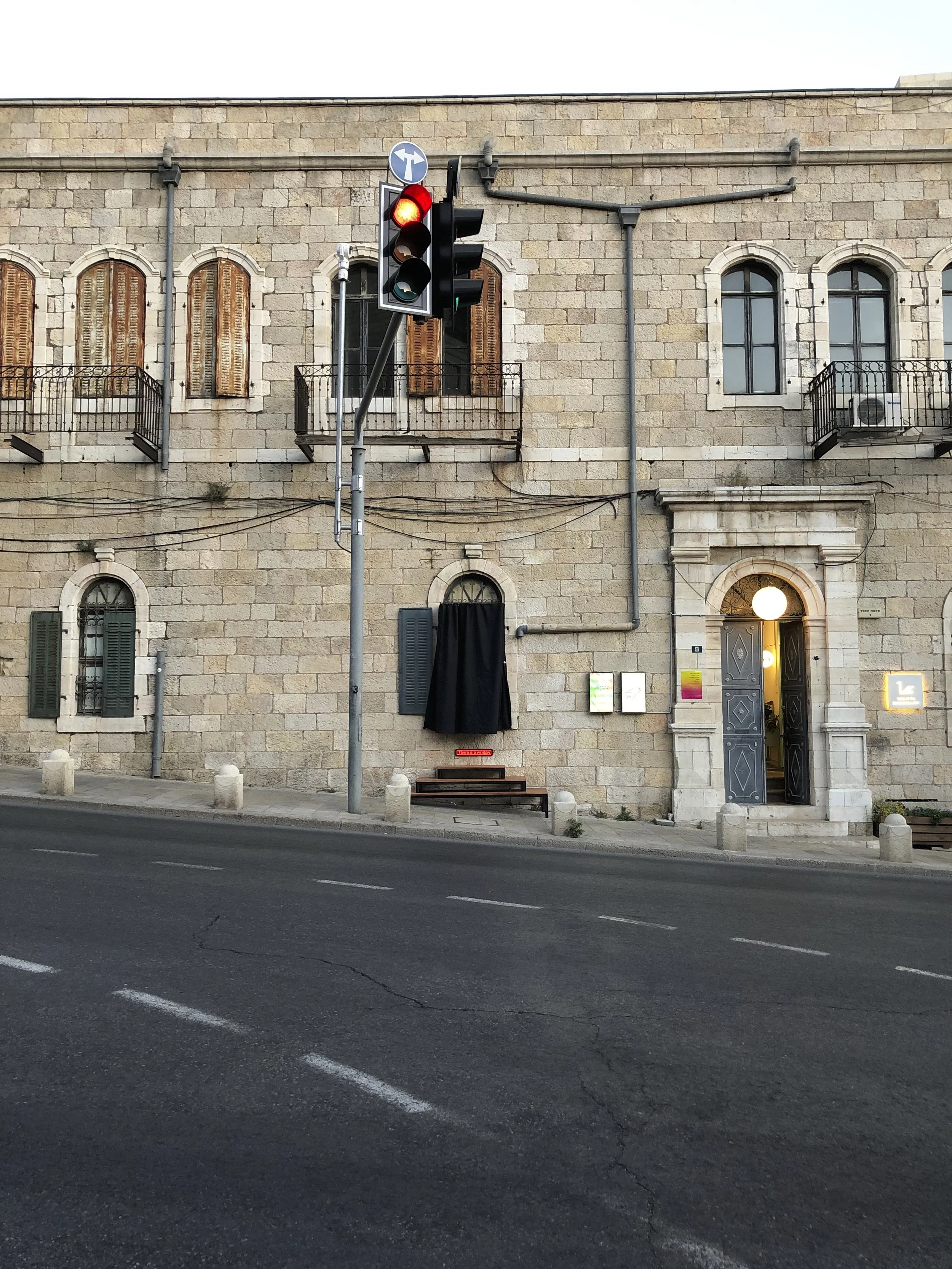 A stone building with arched windows and balcony doors, some with wooden shutters. A traffic light showing red is in the foreground. There is an illuminated doorway on the right side of the building. A black curtain covers a window or doorway in the