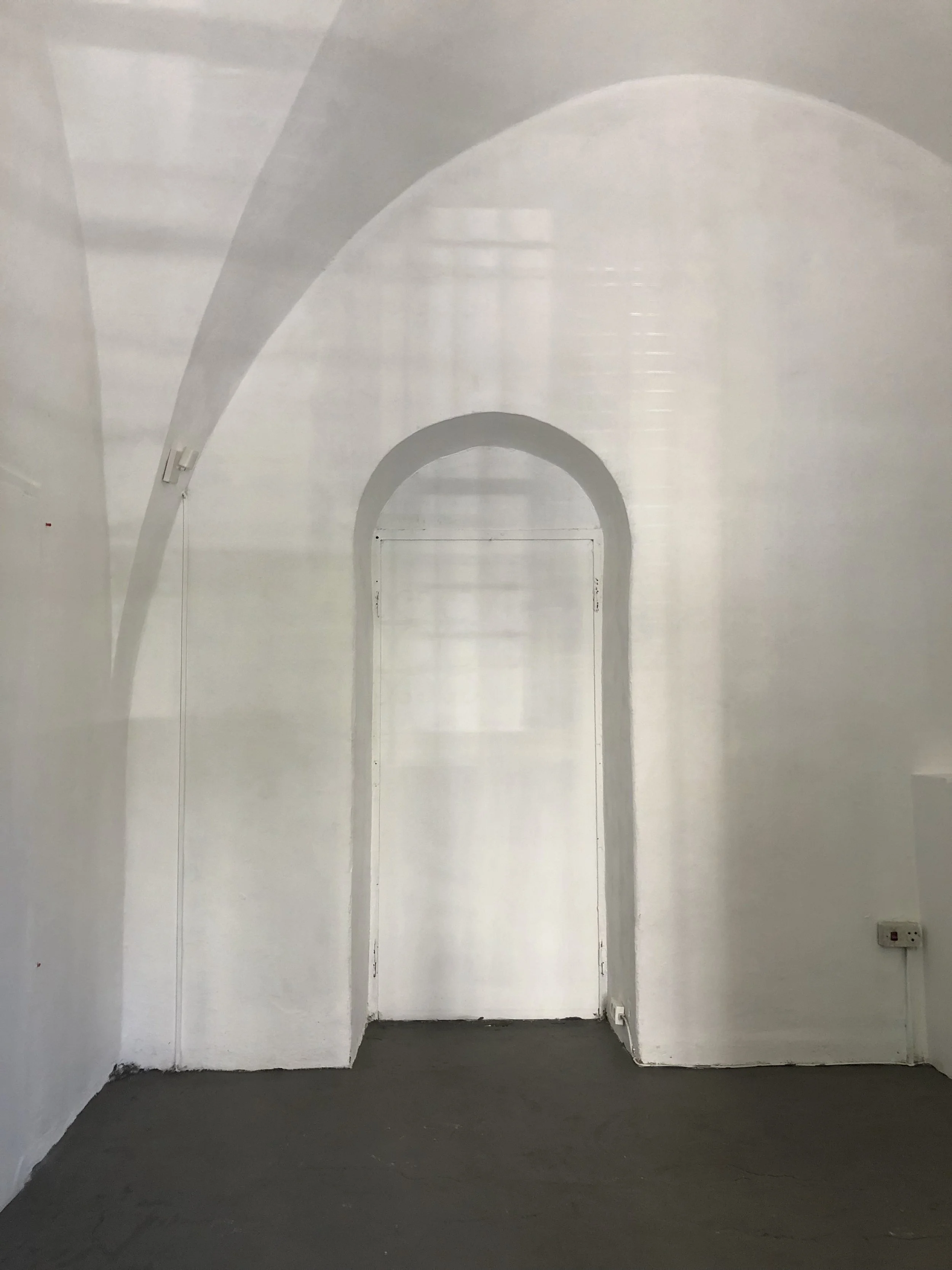 Empty white room with an arched doorway, gray floor, and a power outlet on the wall.