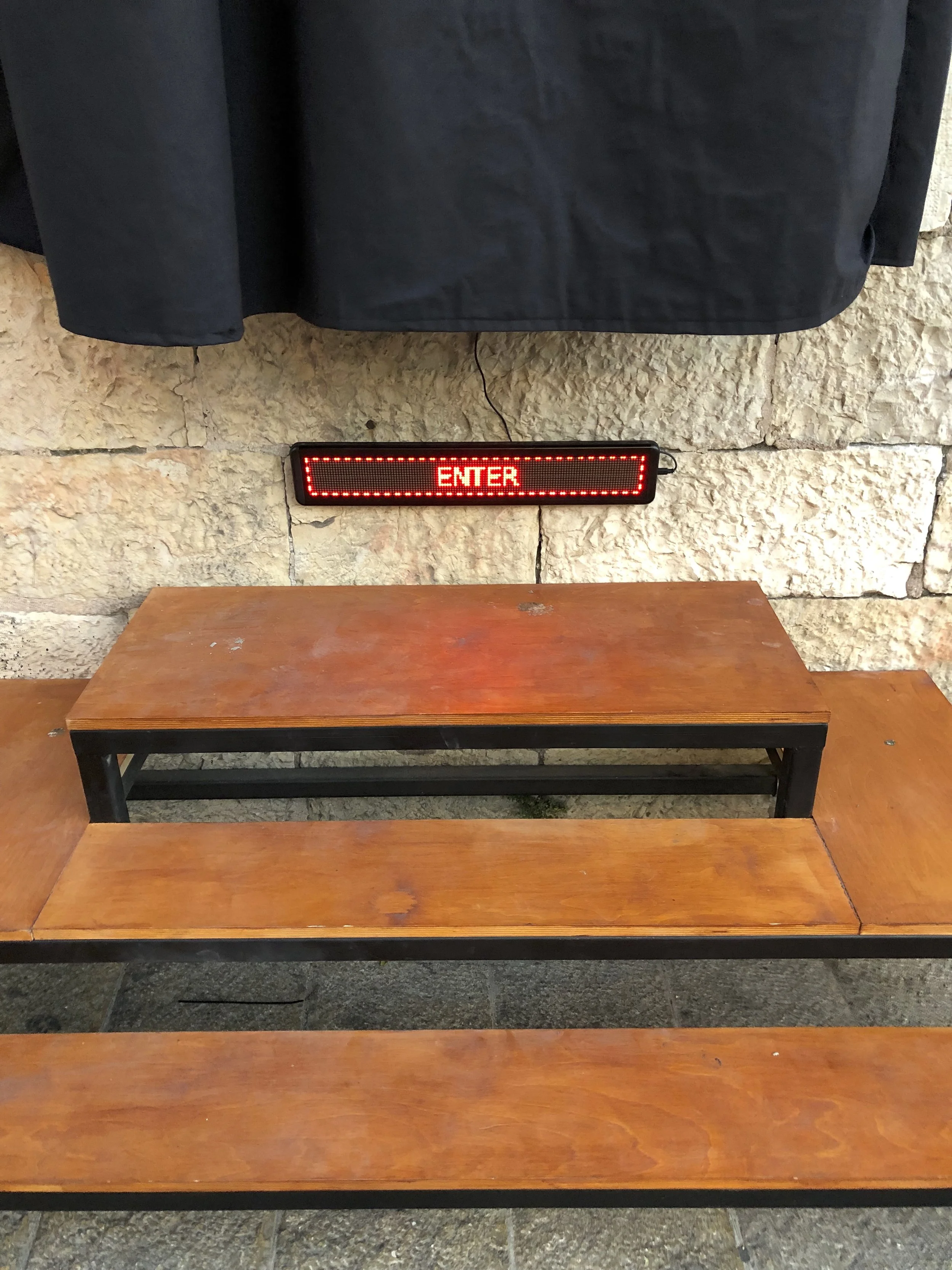 A wooden and metal bench in front of a stone wall with a black coat hanging above, and a red digital sign displaying the word 'ENTER' on the wall.