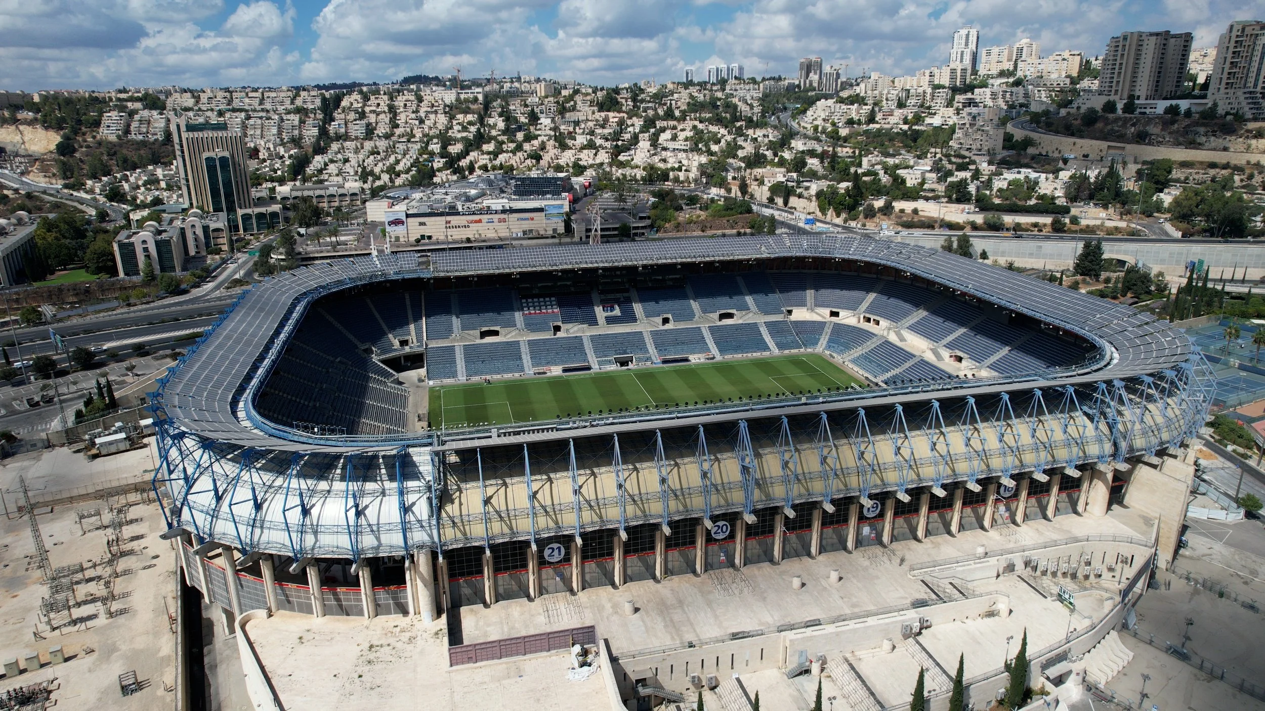 An aerial view of a large, modern outdoor stadium with a grass field and empty seating sections, surrounded by city buildings and roads.