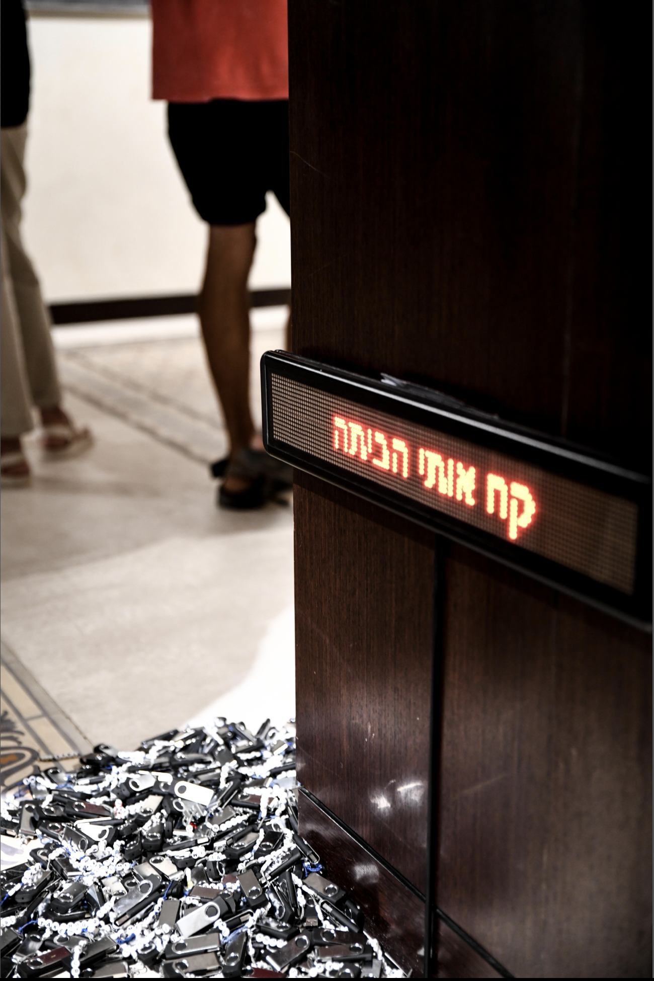 A pile of silver keys on the floor next to a wooden podium with a red electronic sign, in a room with beige carpet and people standing nearby.