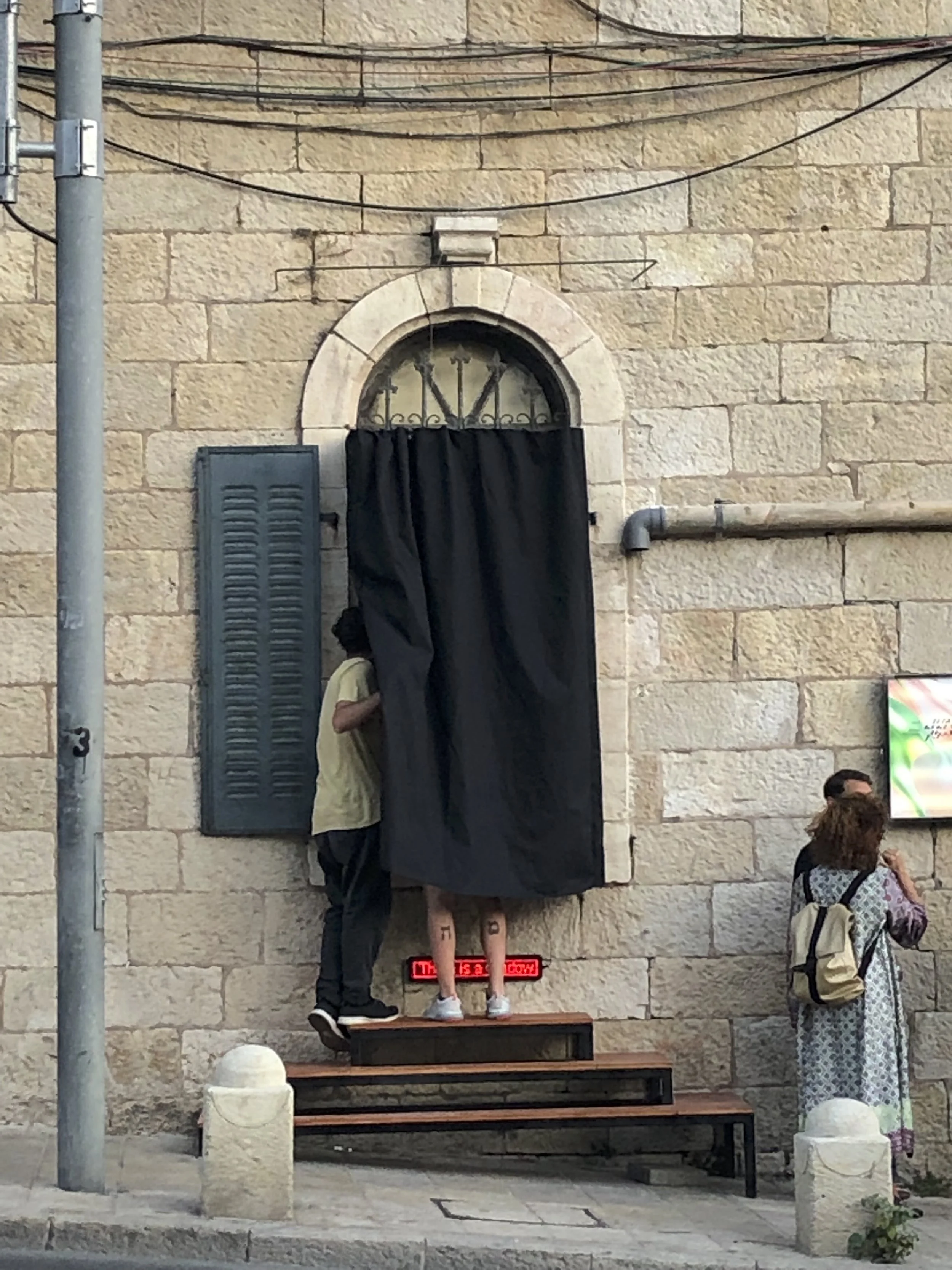 Two people stand on stairs outside a stone building, one with their upper body covered by a black curtain, the other with legs visible showing tattoos, next to a blue shutter. A woman is standing nearby with a backpack, looking at a screen on the wal