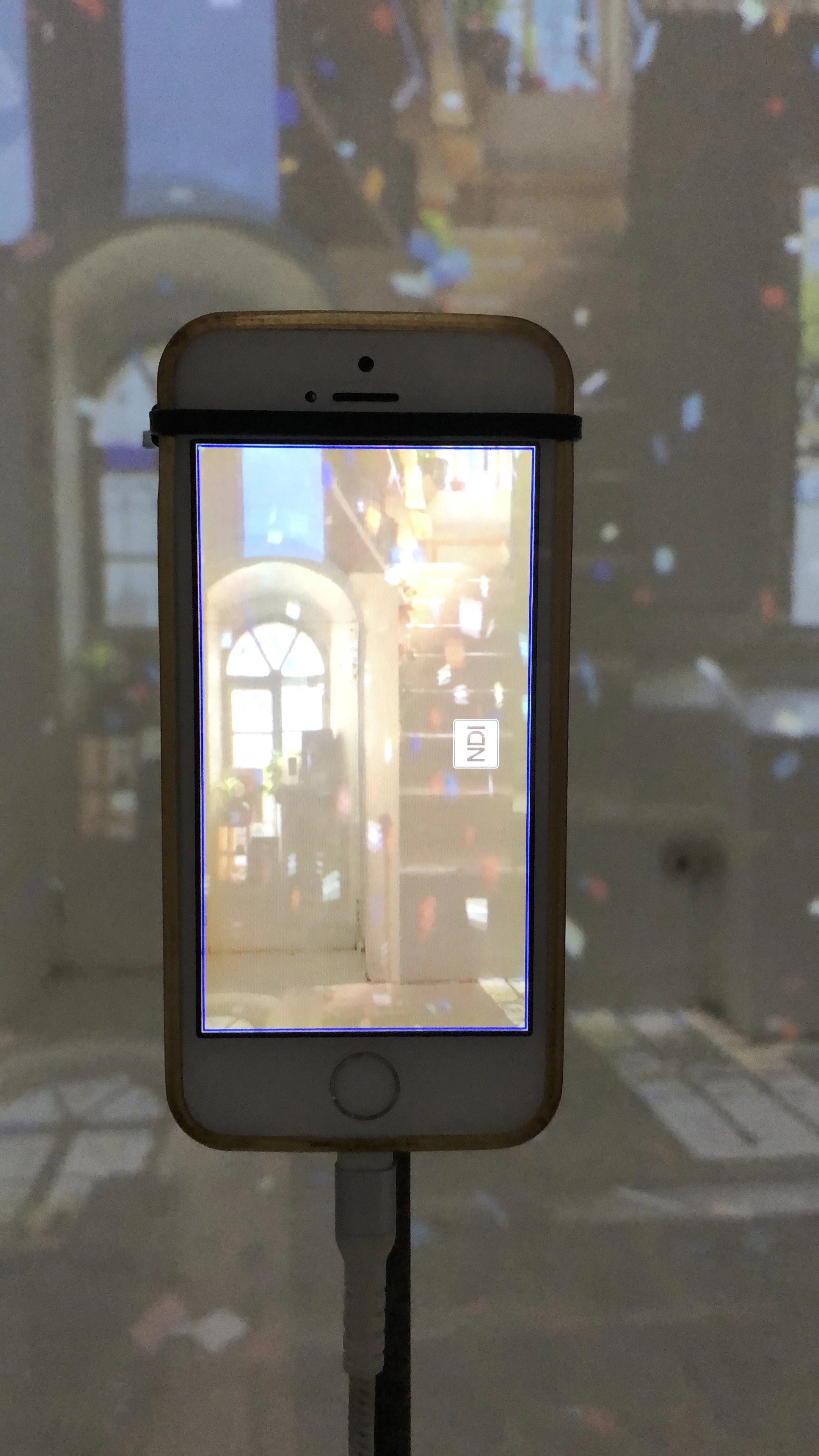 A smartphone displaying a photo of a room with a window, plants, and people, with a projection of the same room on the wall behind it.