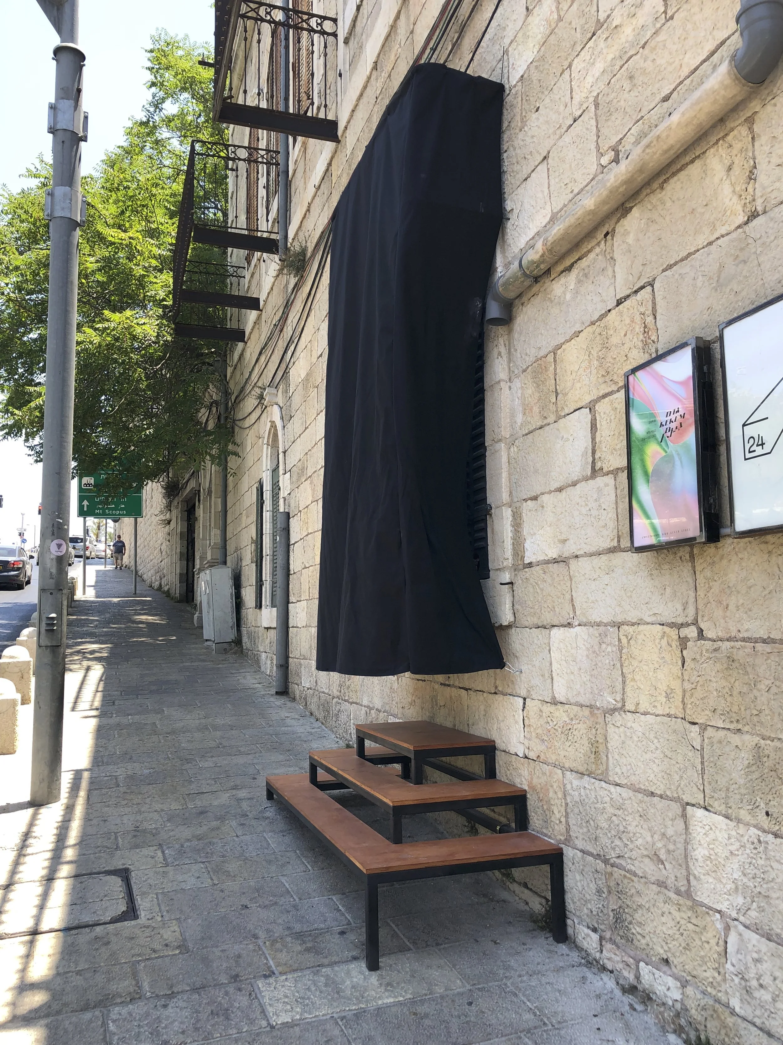Empty outdoor sidewalk with a stone wall, a black cloth covering a window or object, a small wooden and metal bench, and greenery in the background.