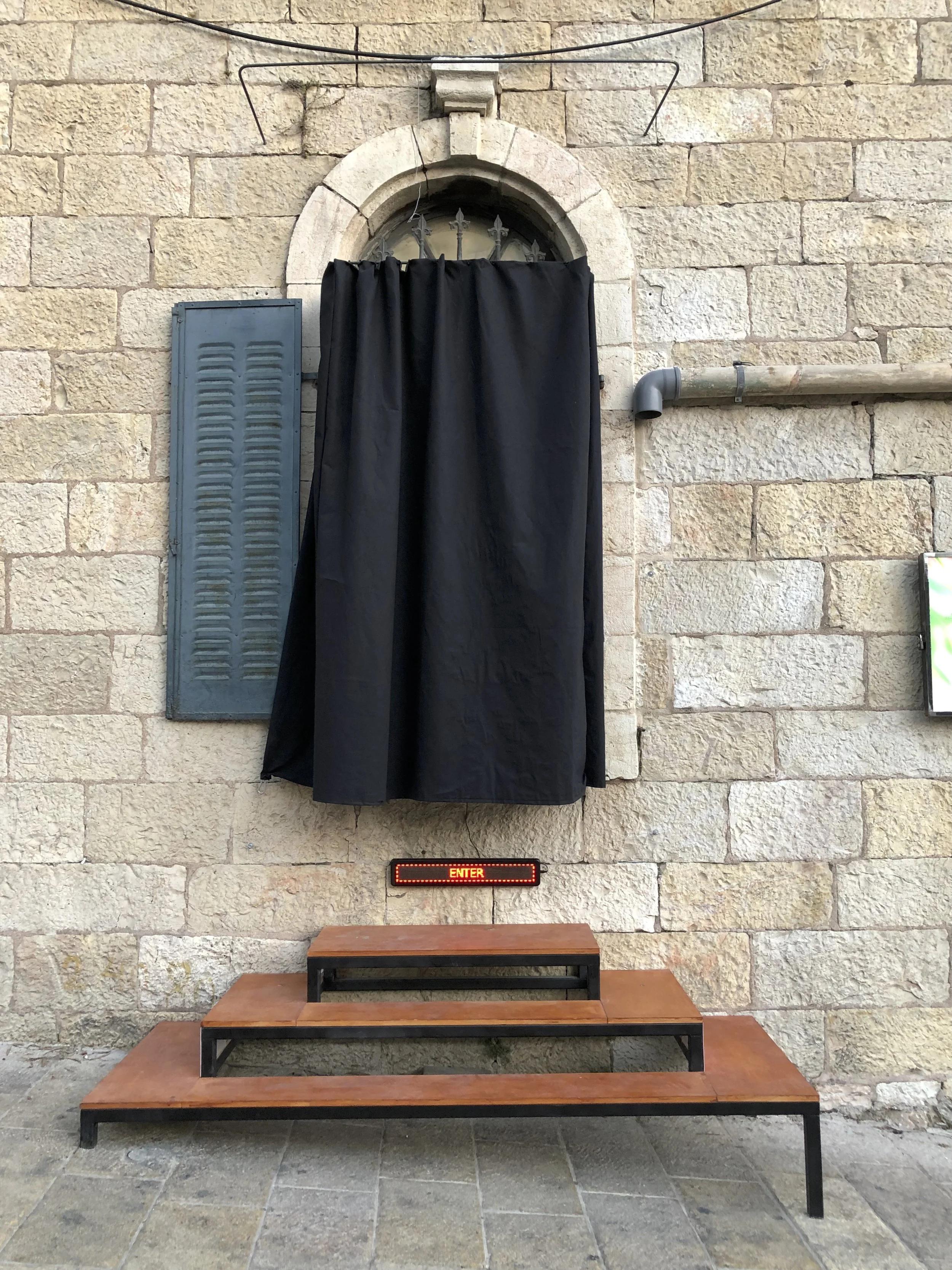 Outdoor scene showing a stone wall with a small arched window that has a black curtain hanging in front of it. To the left is a blue shutter, and on the right is a gray pipe. In front, there are three wooden steps with metal frames, and a digital sig