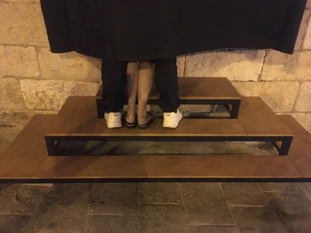 Two people are standing on an elevated wooden platform, with only their legs visible. One person is wearing white sneakers, and the other is wearing black sandals. The background features a stone wall.
