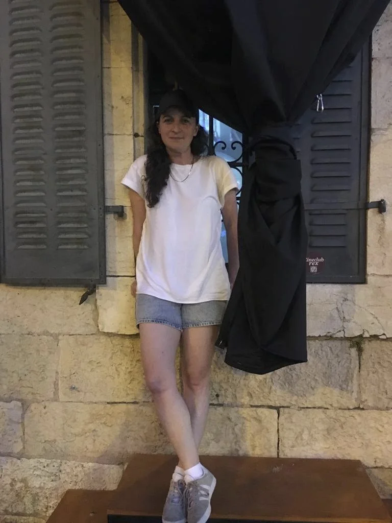 Woman with long dark hair, wearing a white T-shirt, denim shorts, and gray sneakers, standing on a wooden platform in front of a window with black shutters and a black curtain.