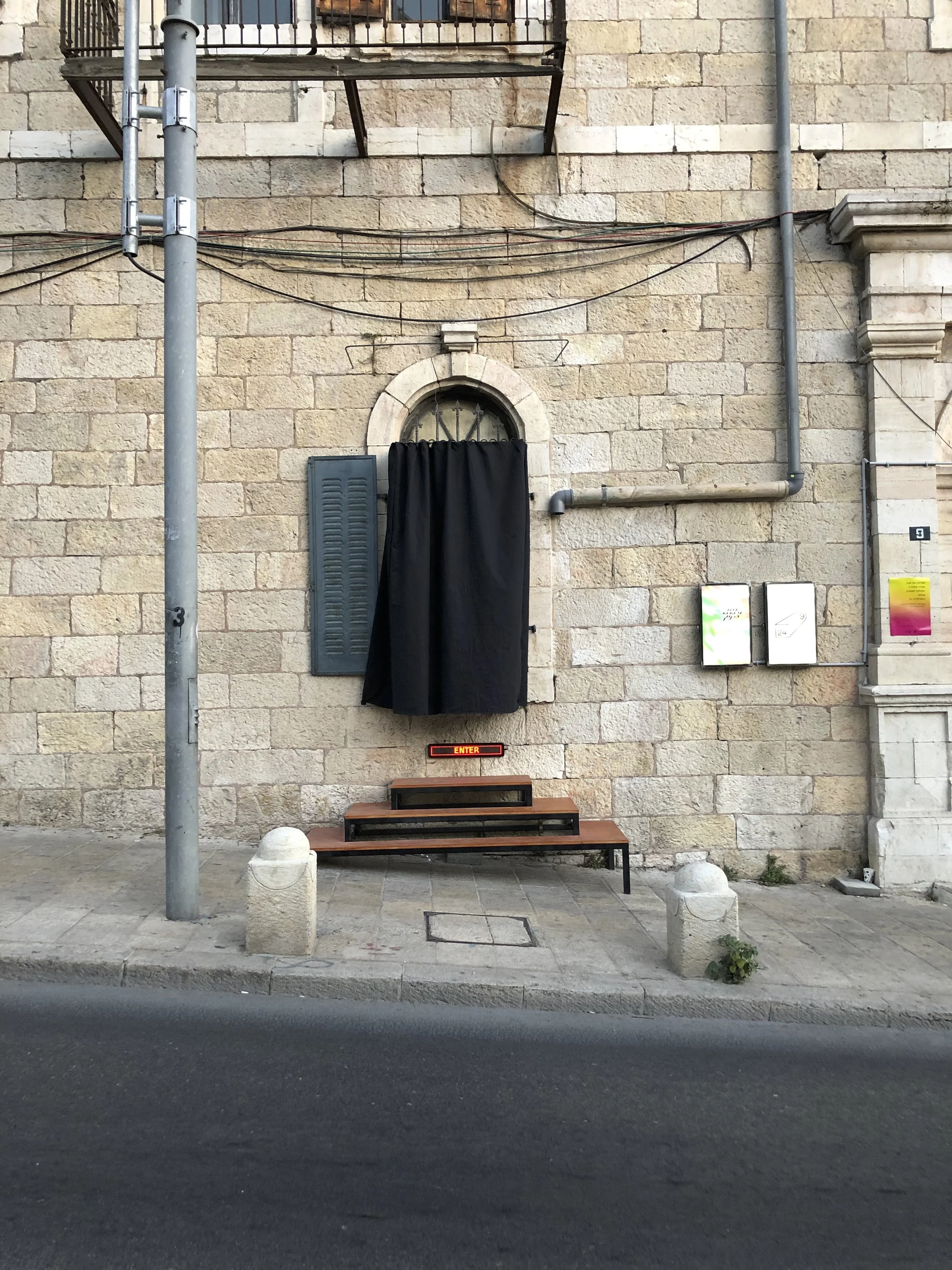 A stone building exterior with a small staircase, black curtain overshadowing a window, and an illuminated 'ENTER' sign at the top of the stairs. Urban street scene.