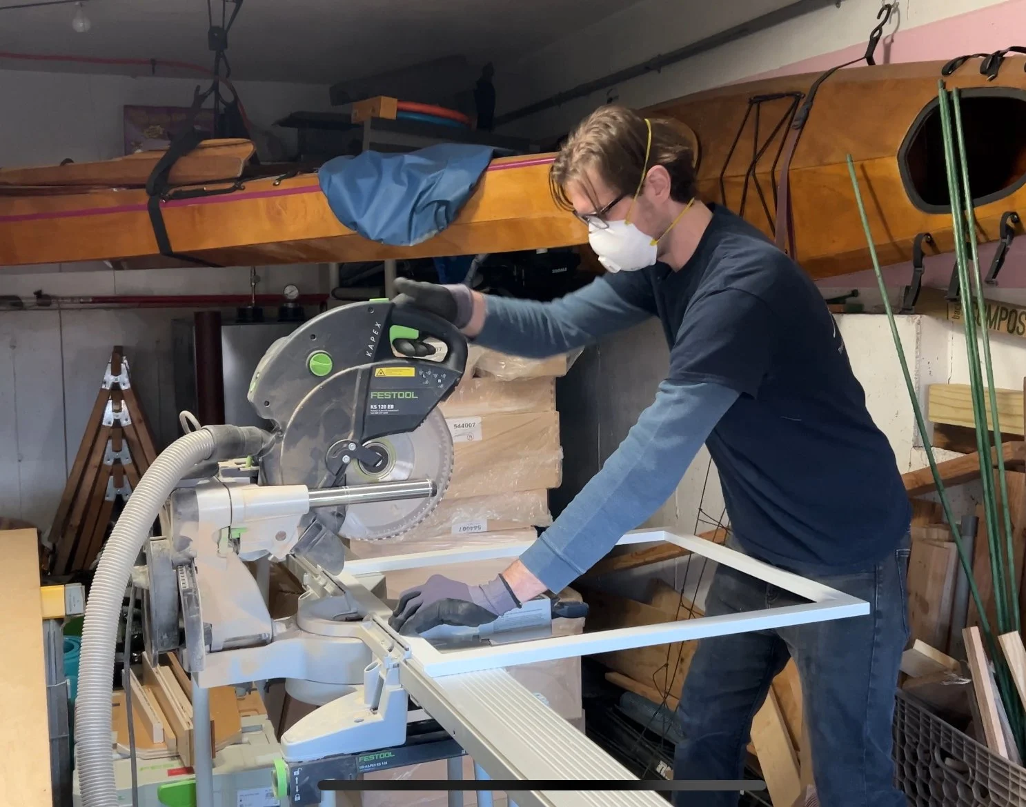 A person wearing safety glasses, a mask, and gloves is using a chop saw in a woodworking workshop. They are cutting a piece of wood, with a kayak and other woodworking tools and materials visible in the background.