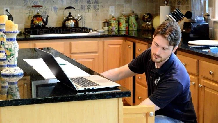 A young man sitting at a kitchen counter with a laptop, working or studying, with kitchen appliances and decorative jars in the background.