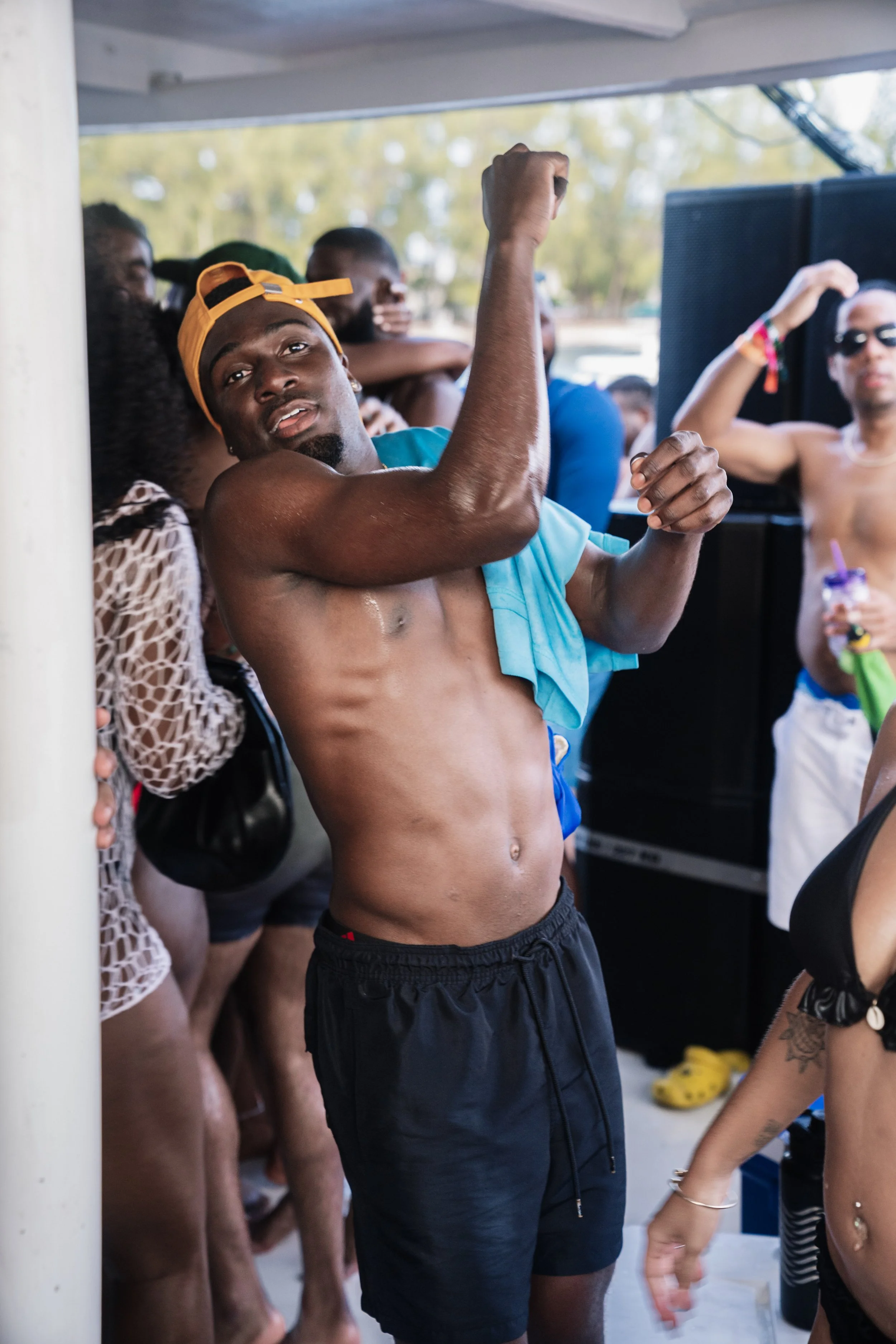 A shirtless man wearing a yellow cap and black shorts dancing at an outdoor party surrounded by other people, some of whom are also shirtless, under a shaded structure.