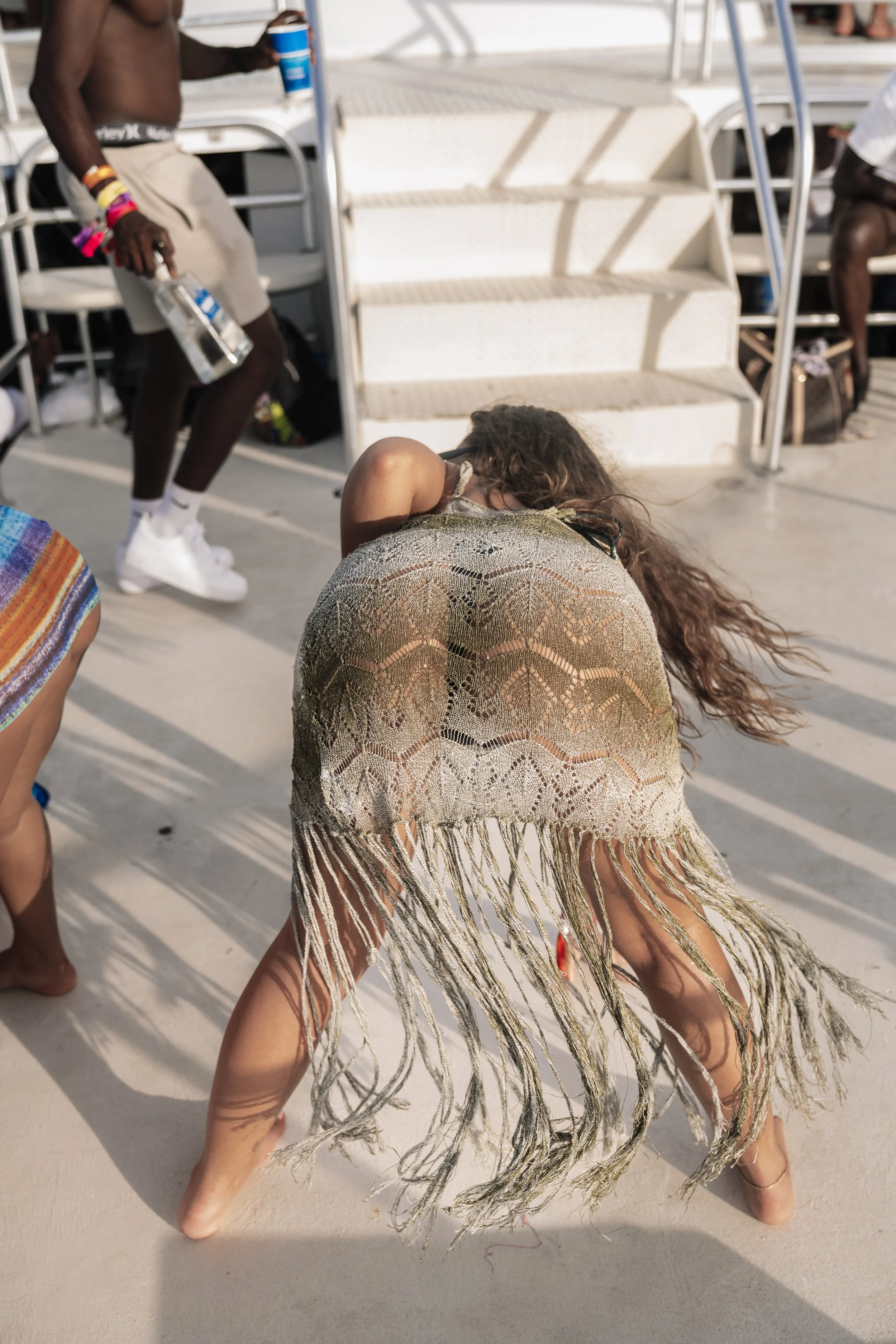 A woman with long hair wearing a fringed, patterned shawl bending over on a sunlit poolside area surrounded by chairs and stairs, with other people sitting in the background.