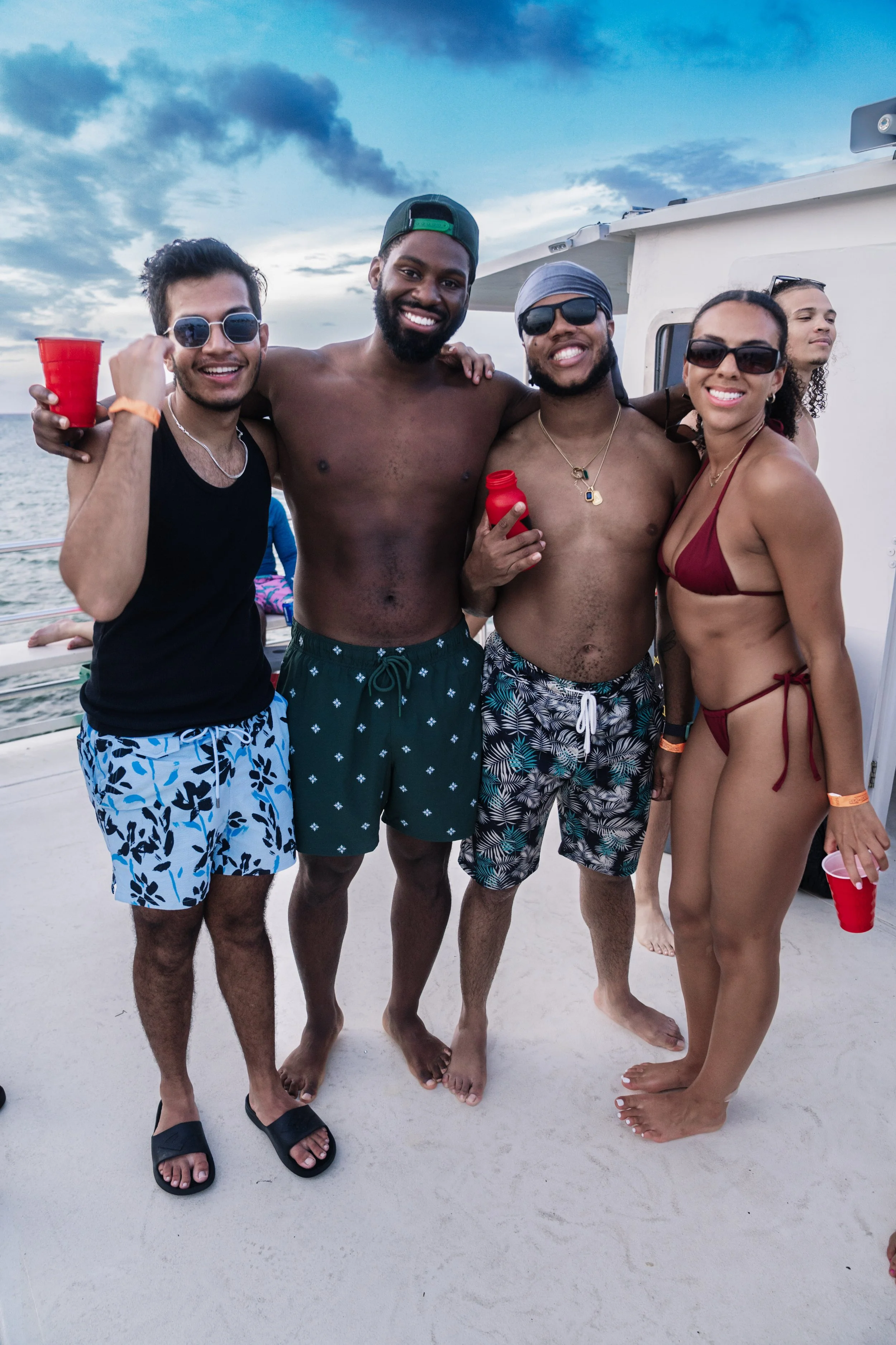 Group of people enjoying a party on a boat, dressed in swimwear, holding red cups, with ocean and sky in the background.