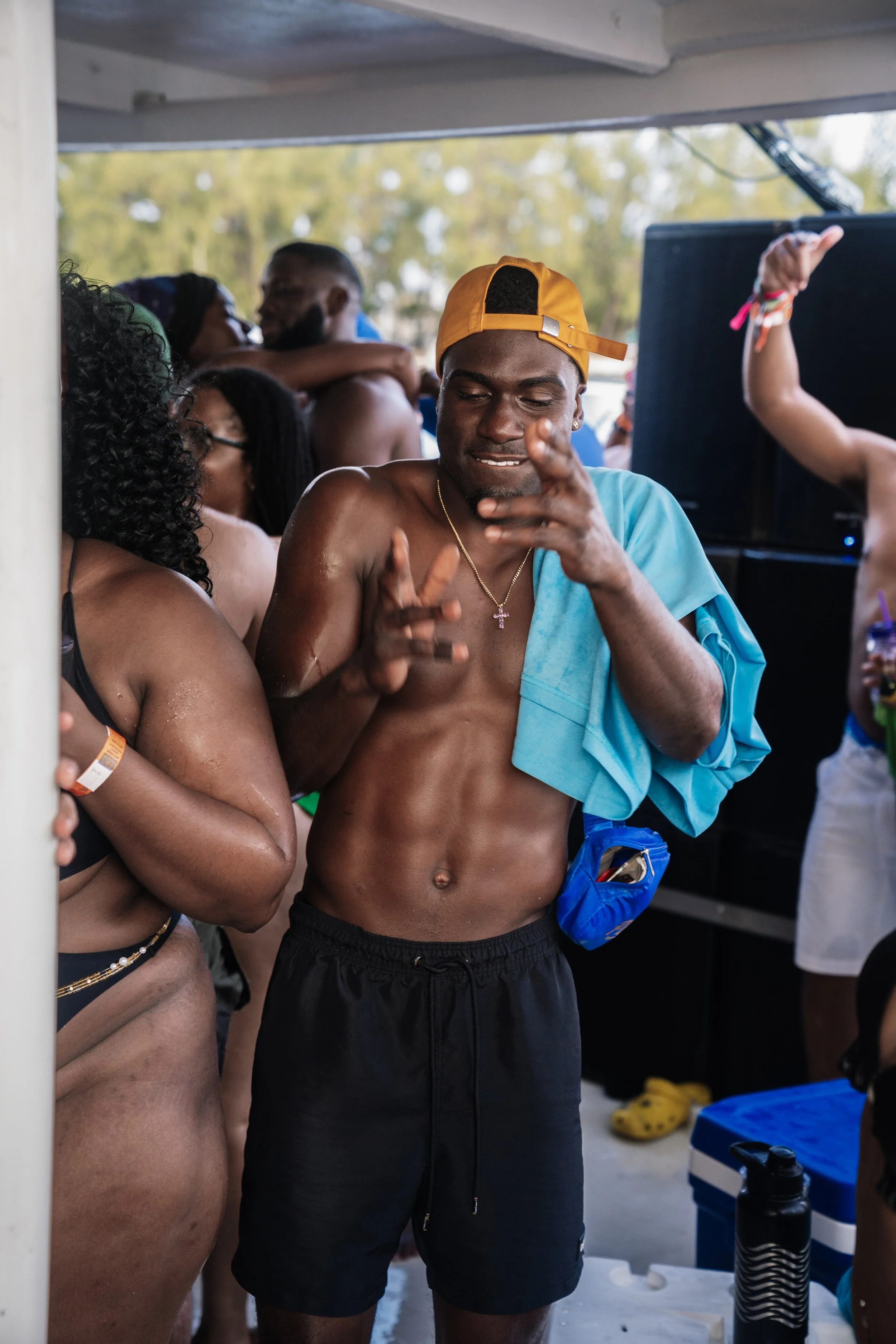 A shirtless man with a yellow cap dancing at a party, surrounded by people in swimsuits under a shelter with trees in the background.