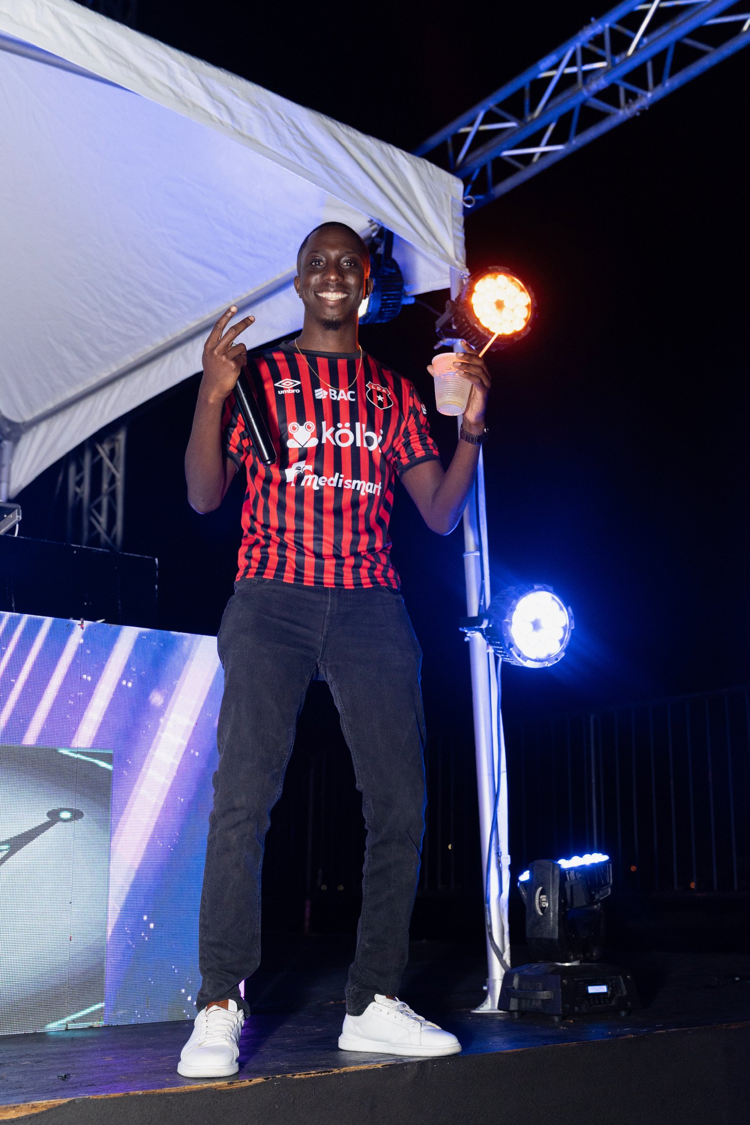 A man in a red and black striped soccer jersey standing on a stage at night, smiling, holding a drink and a lit firework, with stage lights and a large screen in the background.