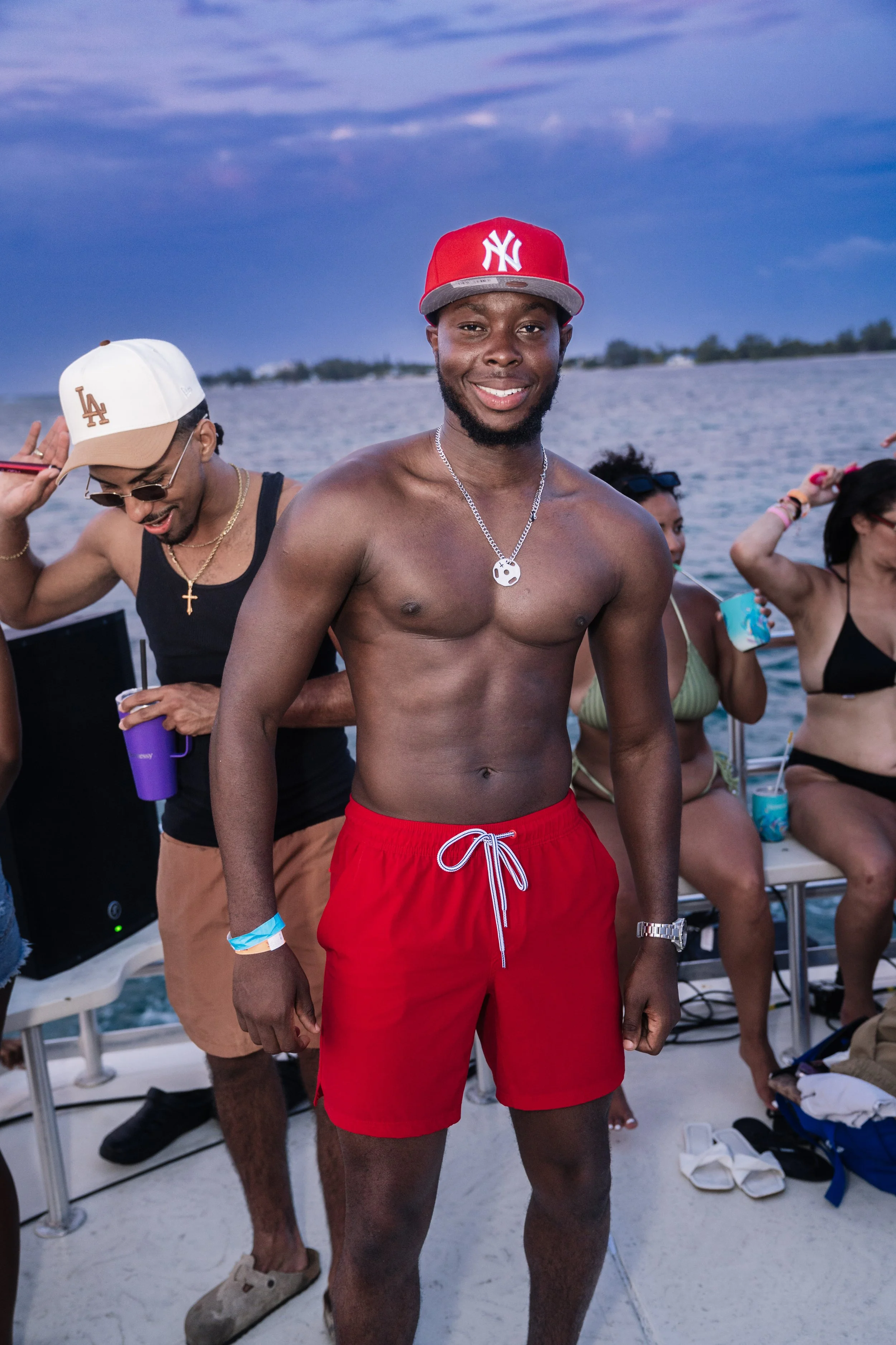 A group of people enjoying a boat party on the water during sunset, with a smiling young man in red swim trunks and a red Yankees cap in the foreground.