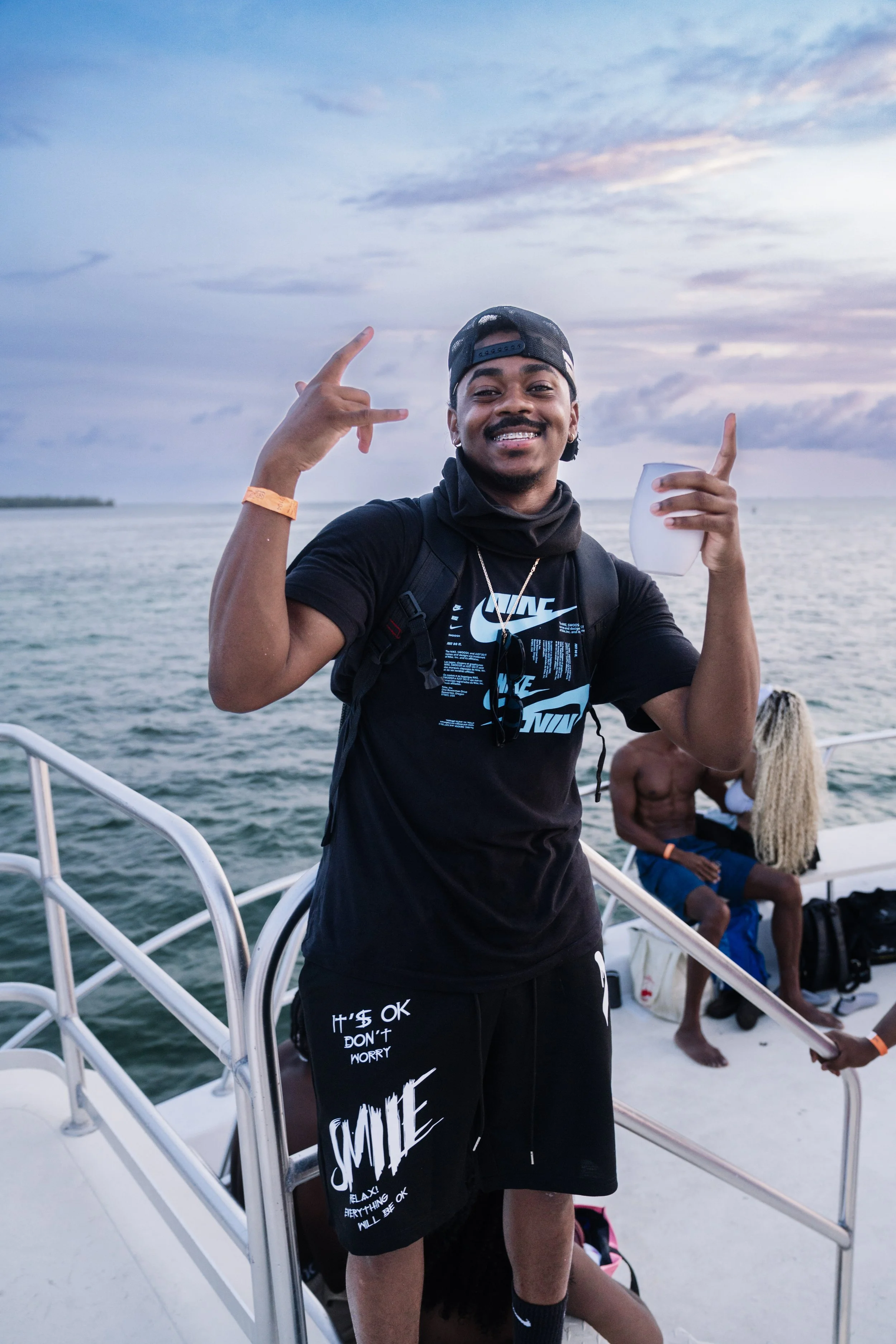 A young man smiling and posing with hand gestures on a boat during sunset with water and a cloudy sky in the background.